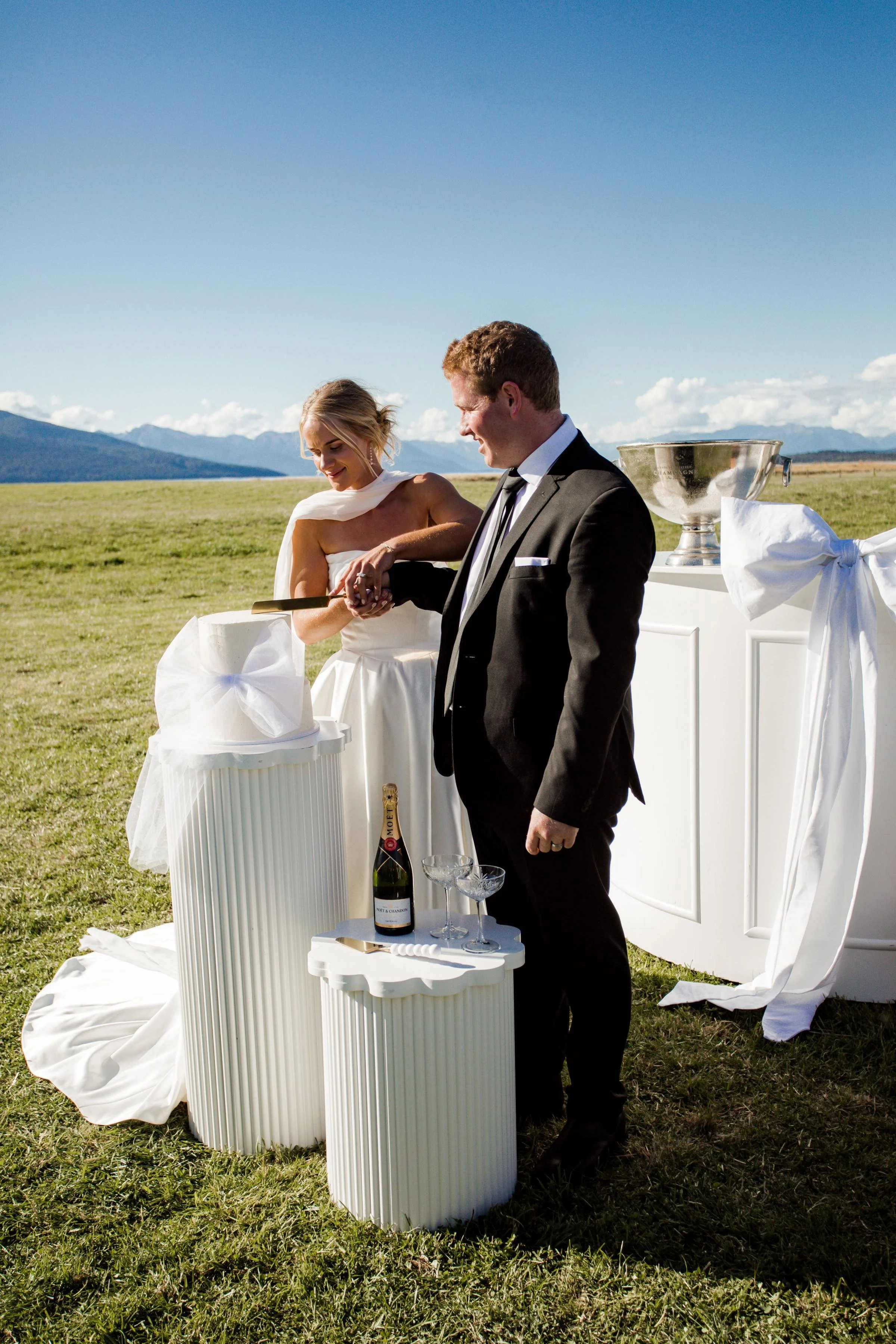 A bride and groom cutting their wedding cake outdoors on a grassy field with mountains in the background. The bride is wearing a white dress and the groom is in a black suit. There are champagne bottles and glasses on a small white table nearby.