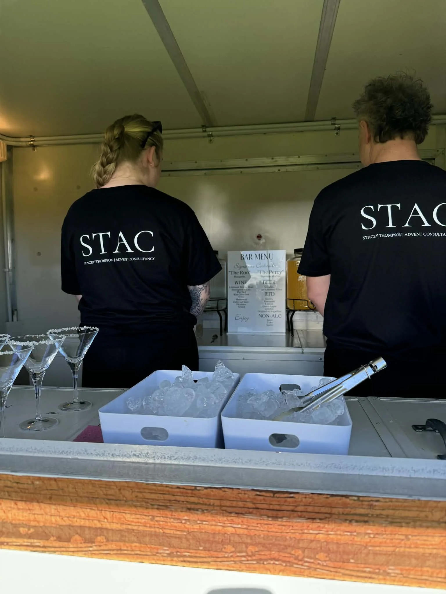 Two people wearing black T-shirts with white text on the back, standing at a bar counter with ice trays and martini glasses, inside a tent or mobile bar setup.