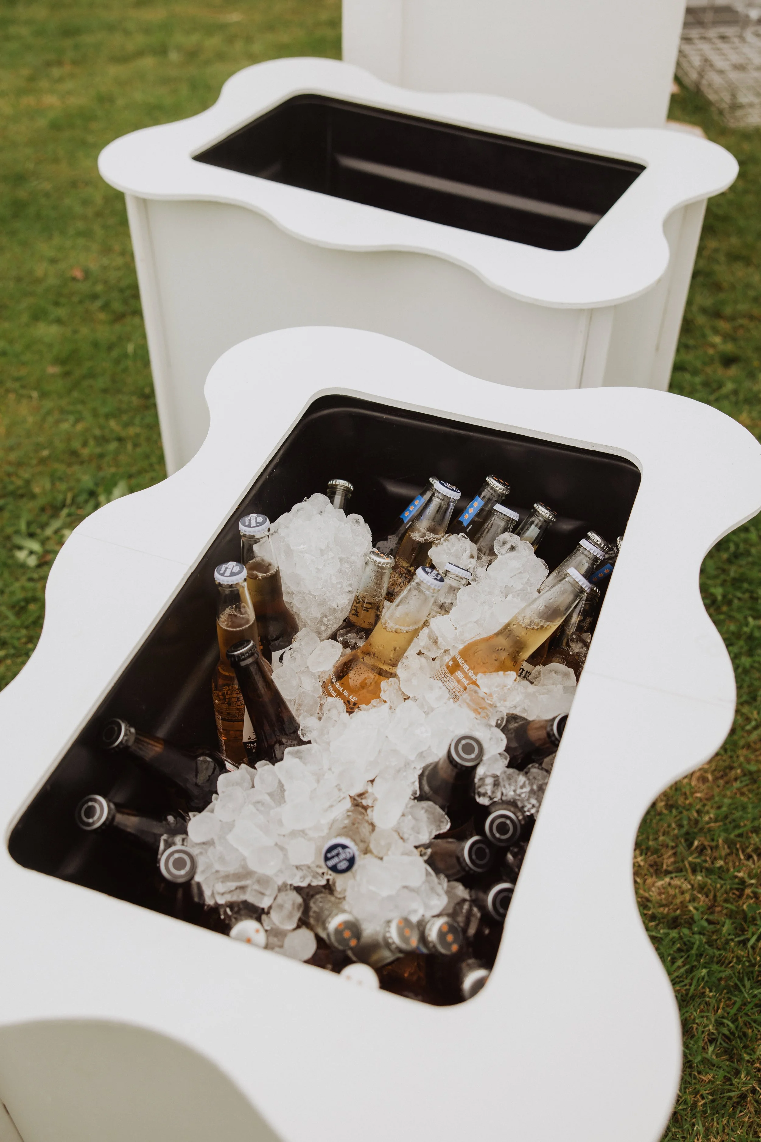 white ice bucket with black interior filled with ice and bottled beverages, outdoors on grassy ground.