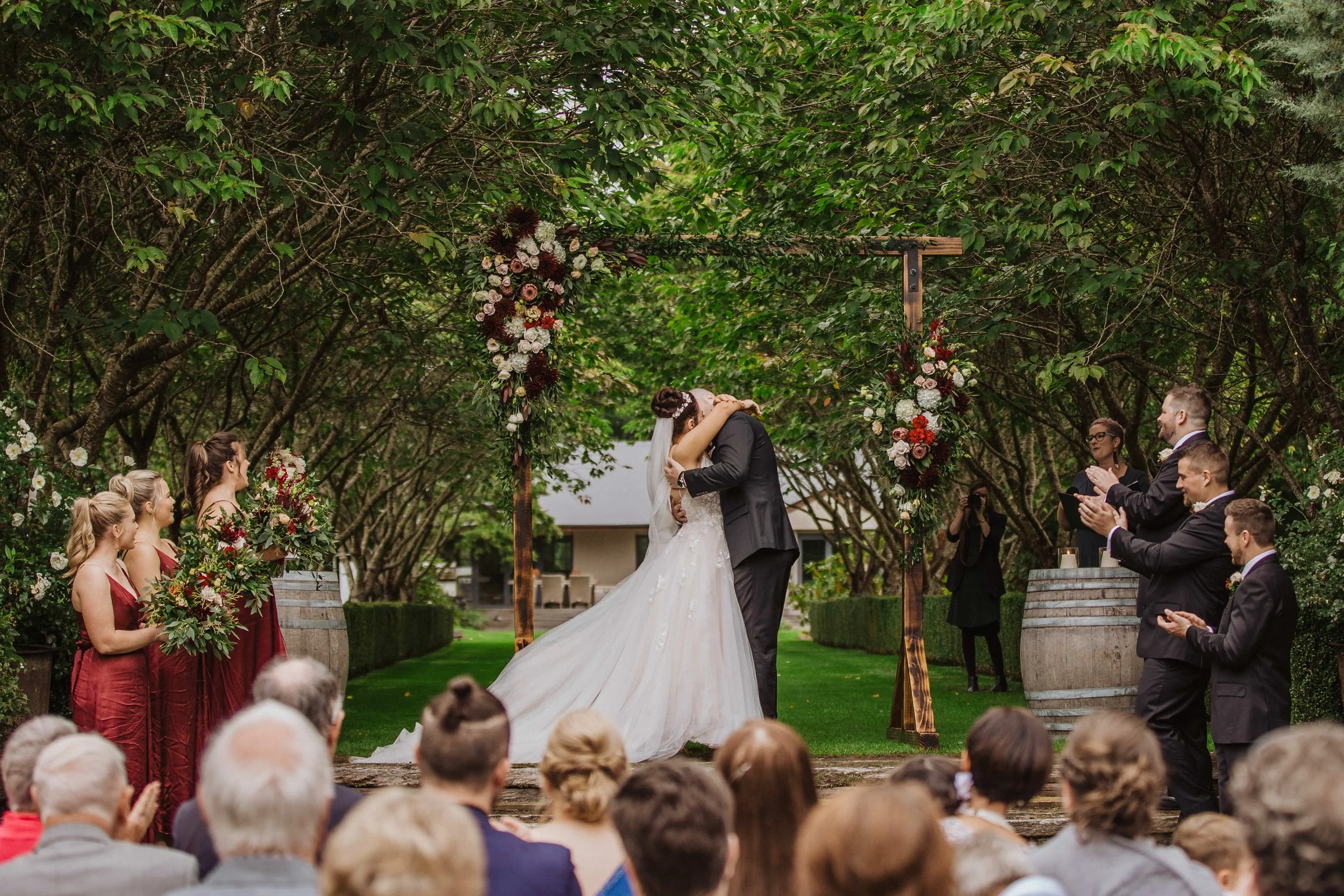 A bride and groom kiss during their outdoor wedding ceremony under a tree arch decorated with flowers, with guests seated and standing around.
