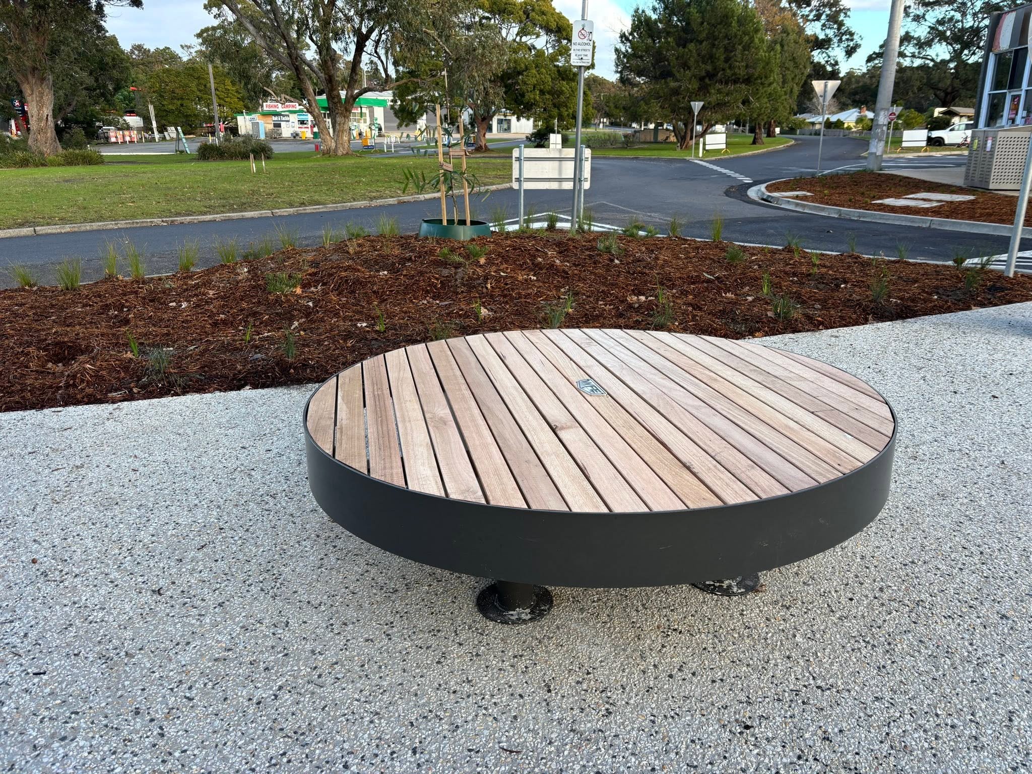 Public park bench with wooden slats and metal frame, surrounded by recently planted landscaping, in a park area with trees, grass, and parking lot in the background.