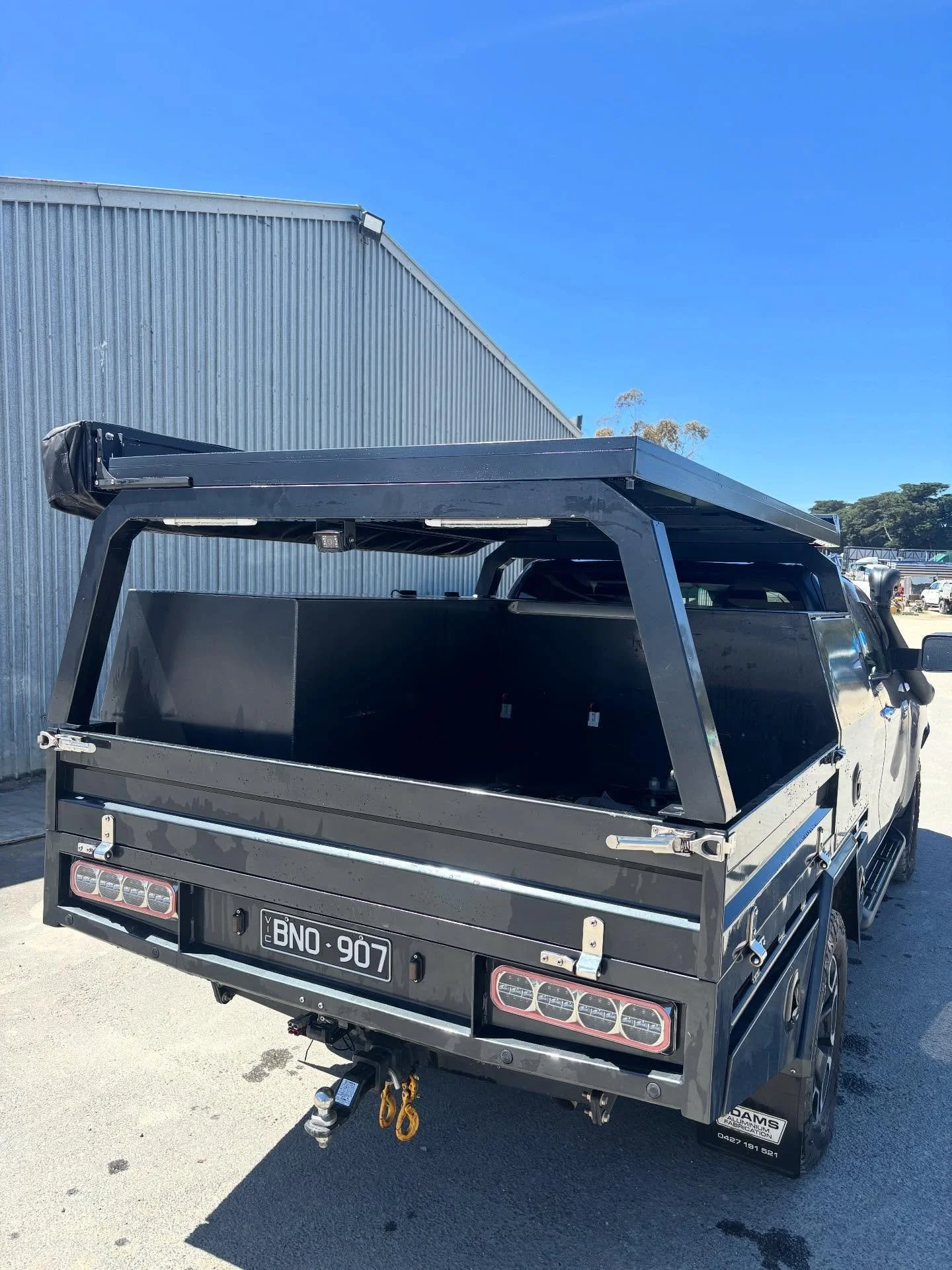 Front view of a black pickup truck with an open cargo bed, parked outdoors under clear blue sky, with a metal building and trees in the background.