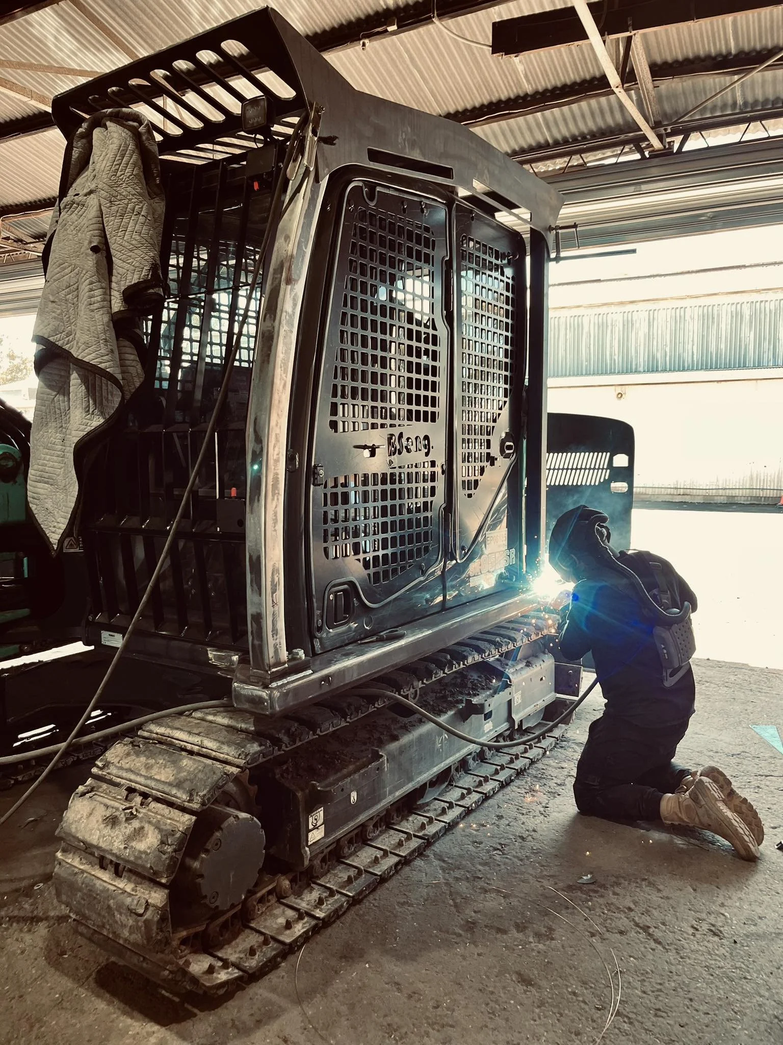 A person kneeling while working on a construction vehicle inside a large warehouse or workshop. The vehicle appears to be a tracked machine with an open cab, and the person is using a tool near the vehicle's side.