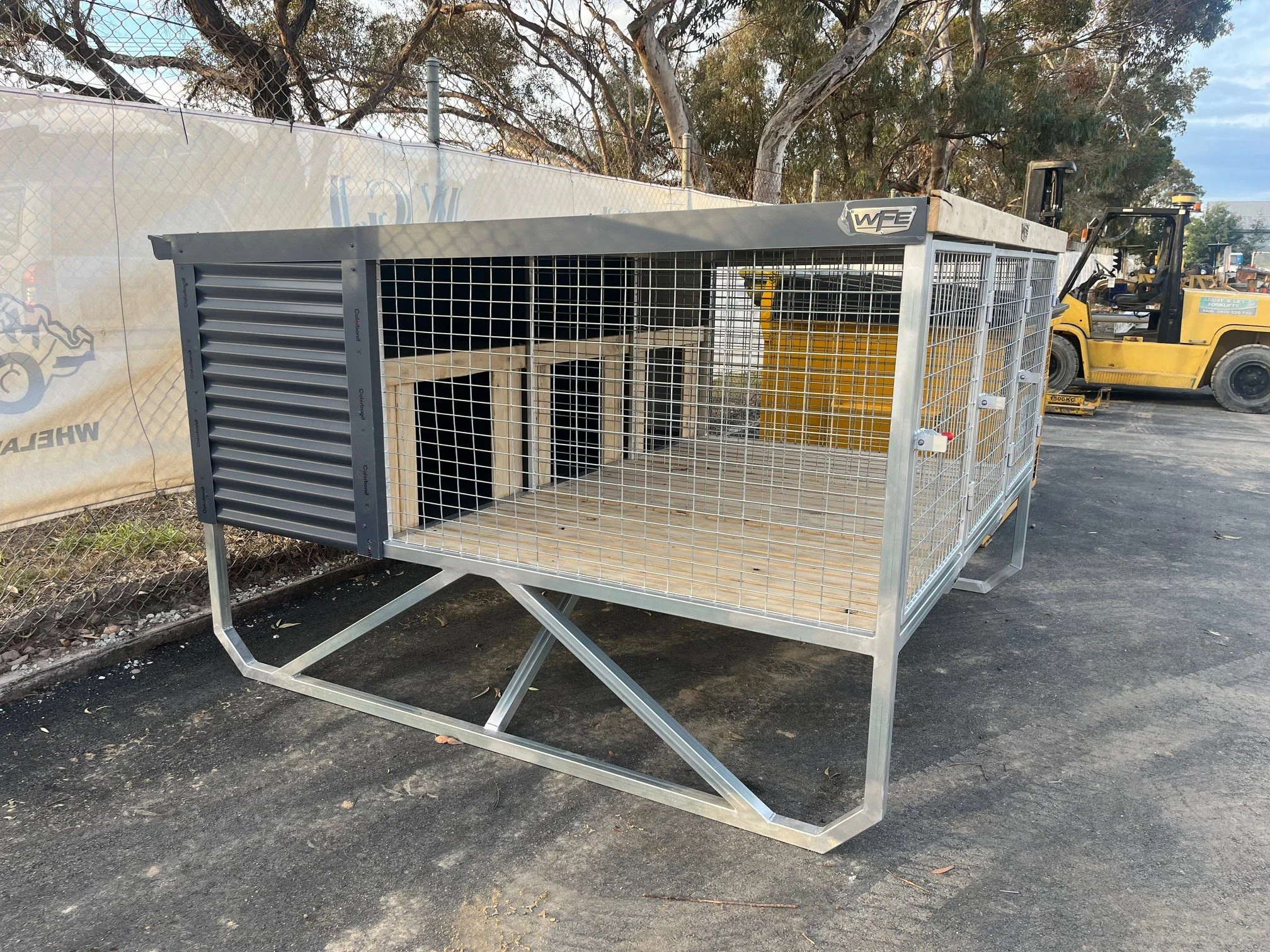 A metal cage on wheels with a wooden doghouse inside, situated outdoors on a paved area near a fence and trees, with construction equipment visible in the background.