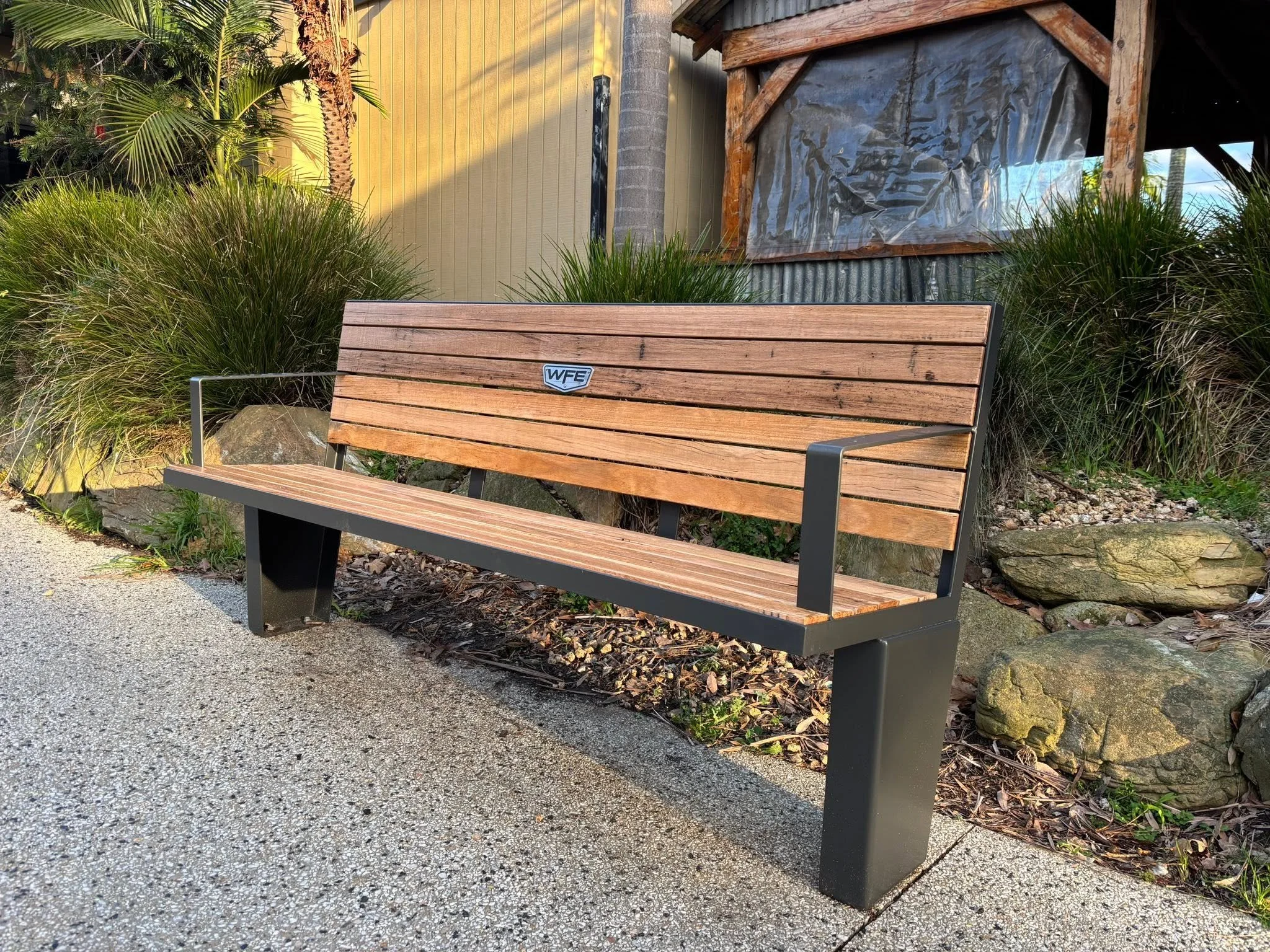 A wooden and metal park bench situated on a gravel pathway with plants and a wooden structure in the background.