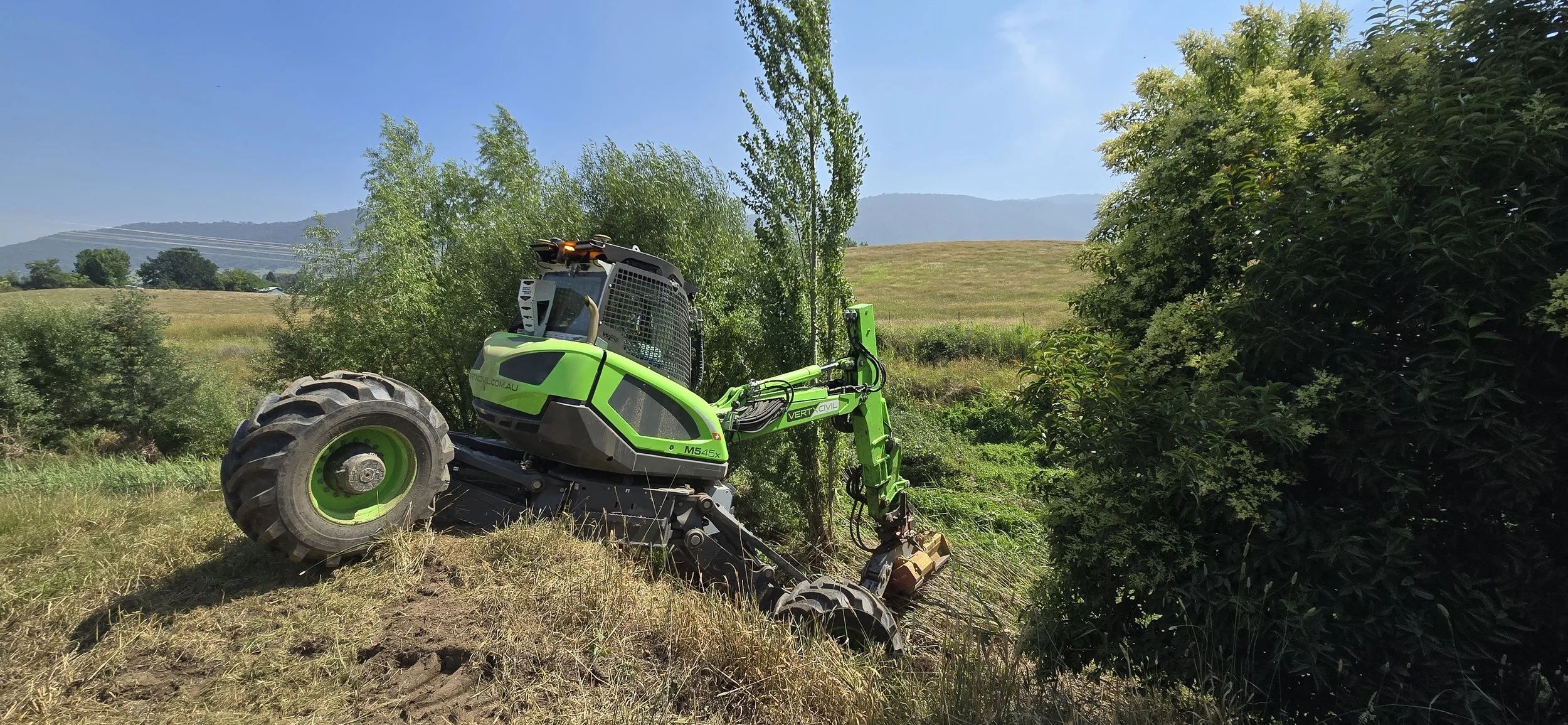 A green robotic forestry machine trimming bushes in a rural landscape with trees, grass, and distant hills under a blue sky.