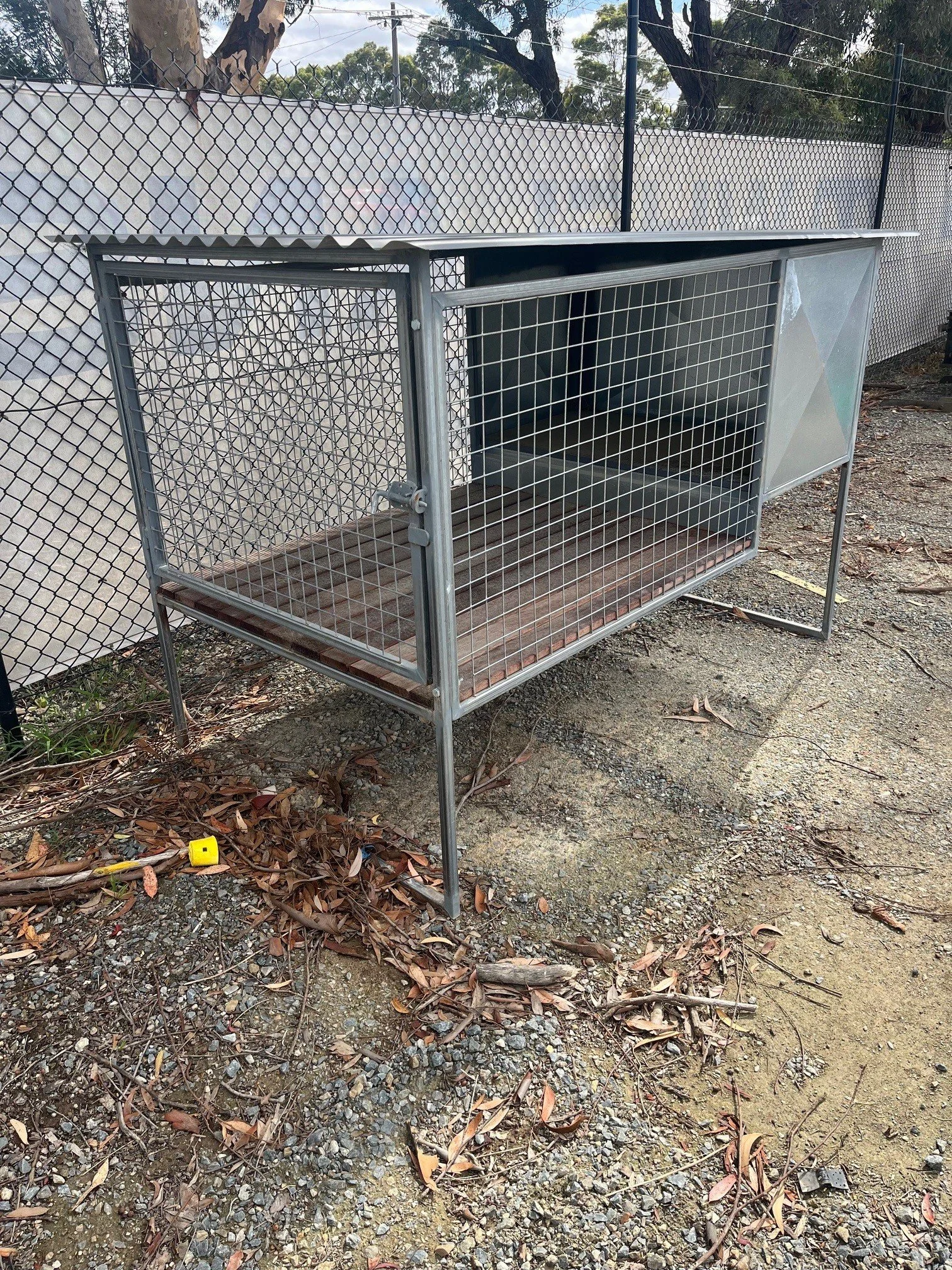 Empty outdoor animal cage with metal frame, wire grid sides, and a sliding door, on rough, leaf-strewn ground with a chain-link fence and trees in the background.