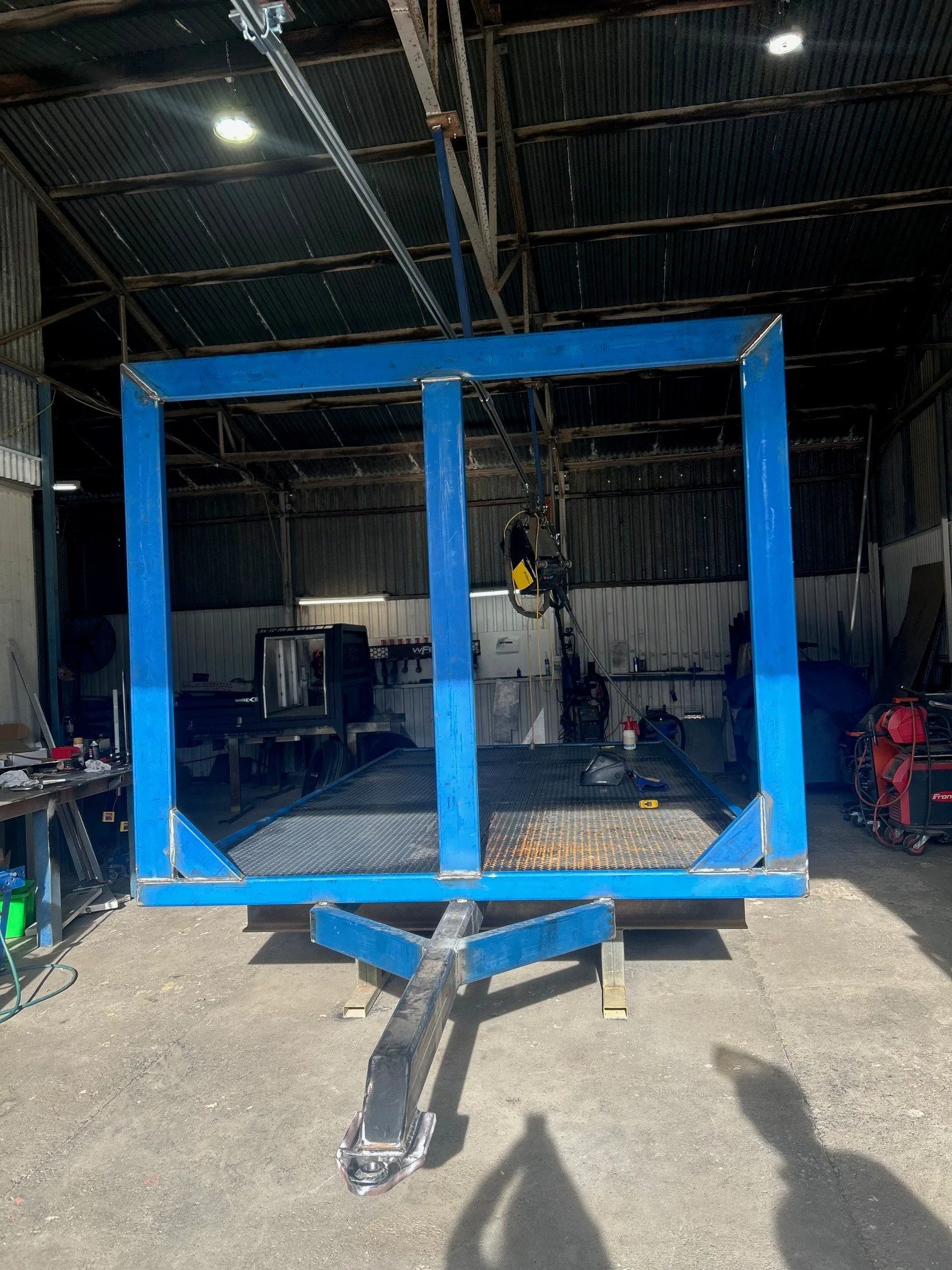 A metal trailer with a blue frame and black surface inside a workshop or garage, with various tools and equipment visible on the sides and background.