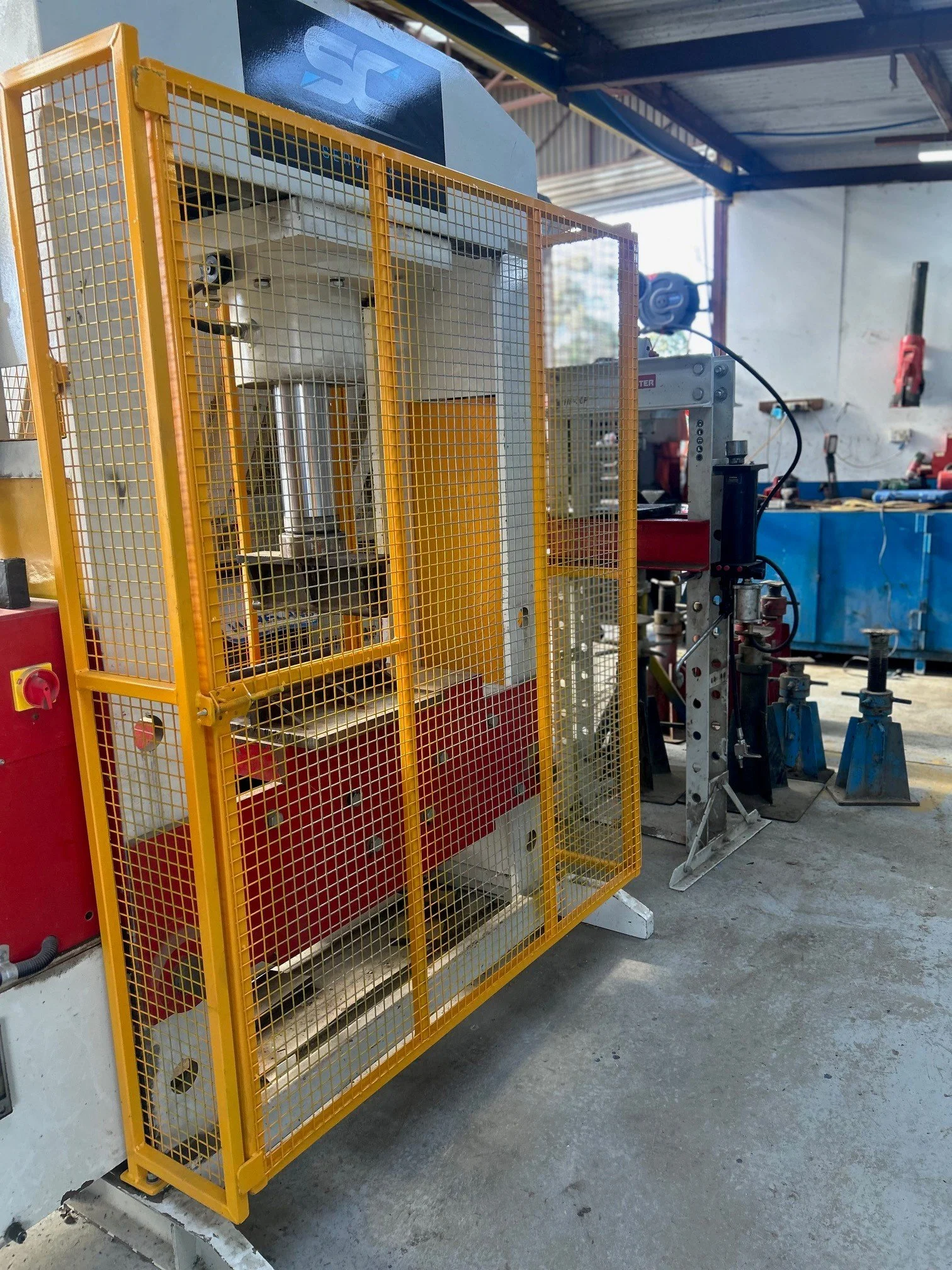 Industrial workshop with machinery enclosed in a yellow safety cage, various tools on a blue workbench, and a white wall with a vertical drill press mounted on it.