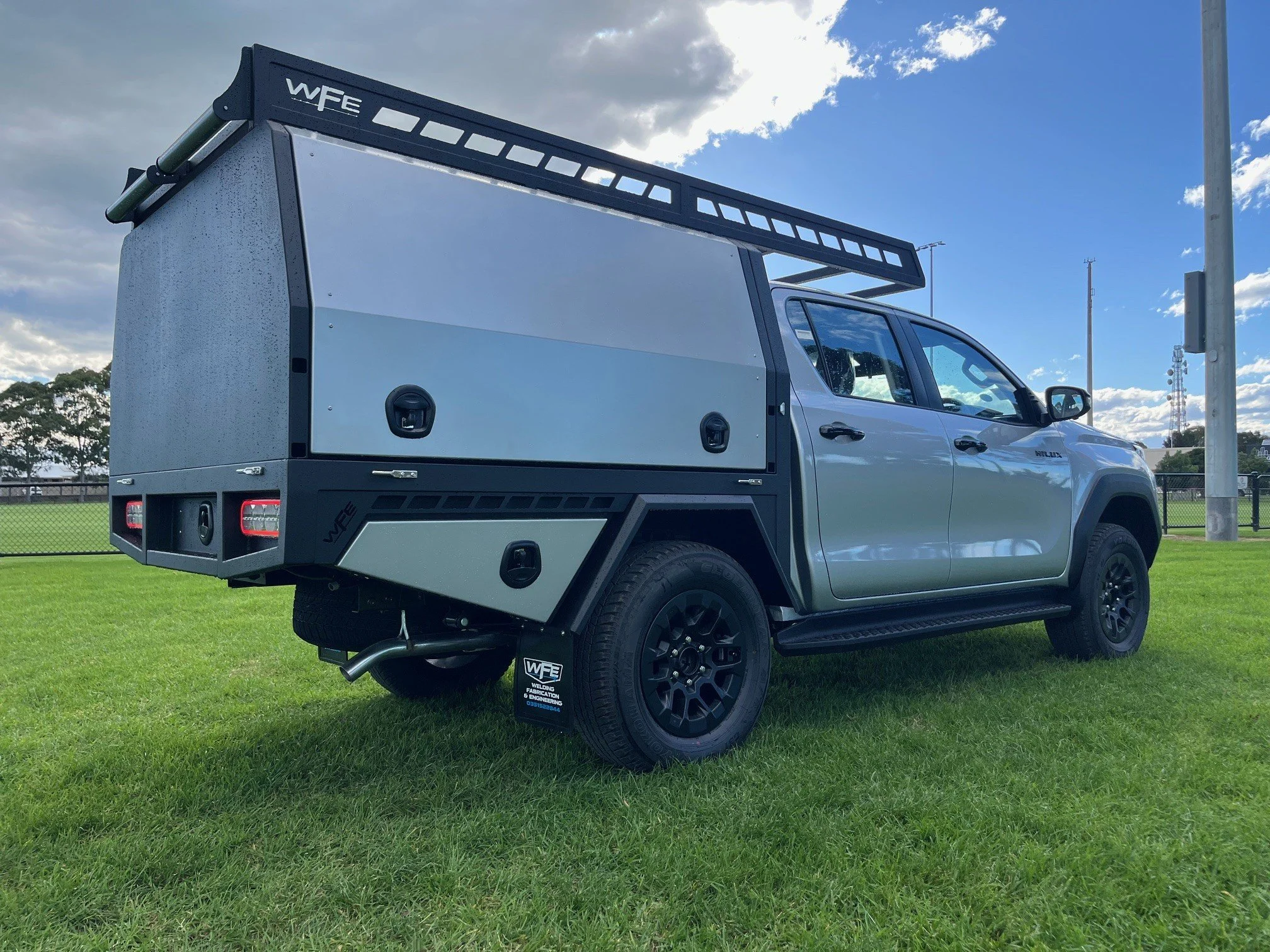 A white utility truck with a black roof rack and storage compartments parked on a grassy field under a partly cloudy sky.