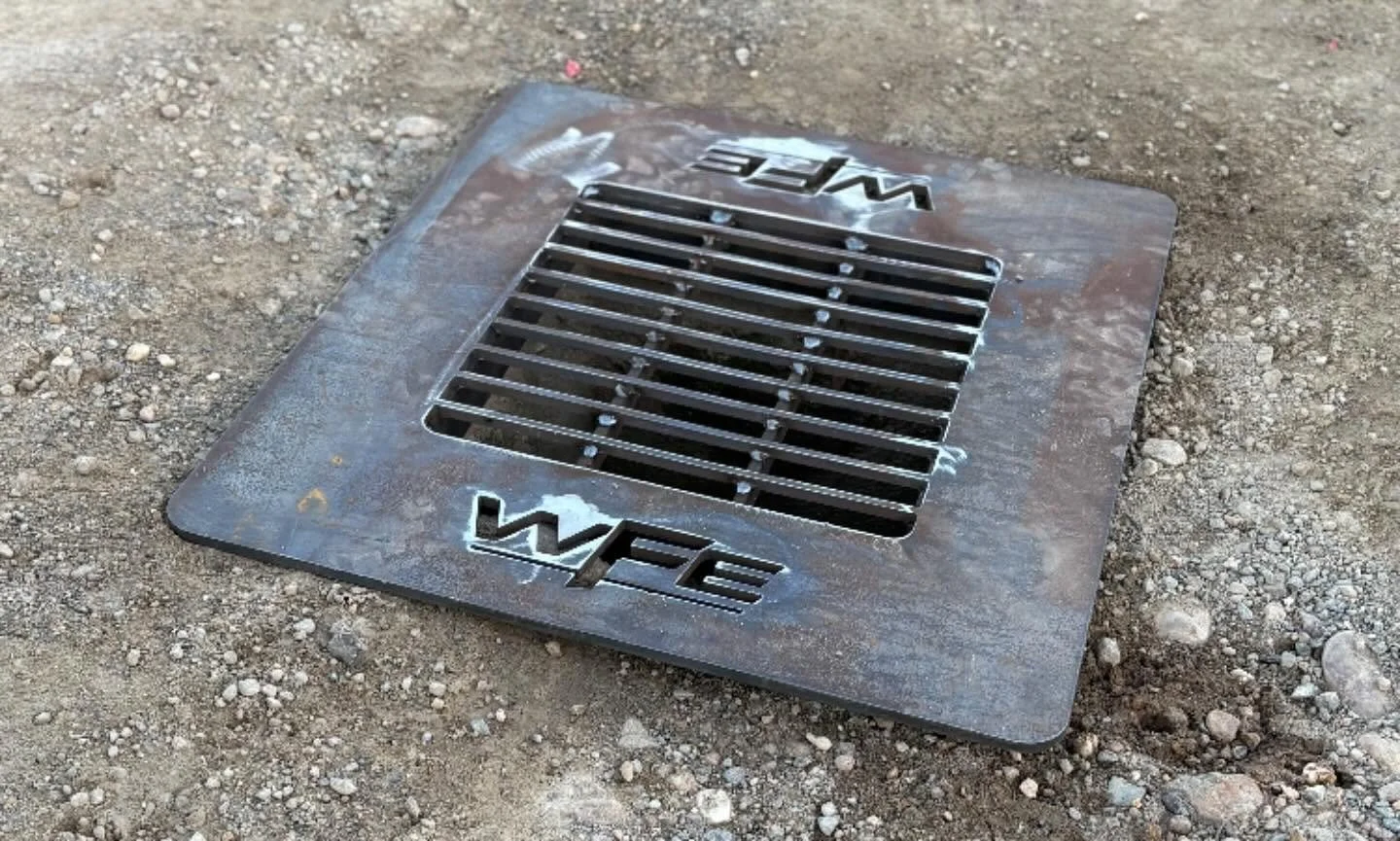 Metal drain cover with the word 'WET' cut out twice, placed on dirt ground.