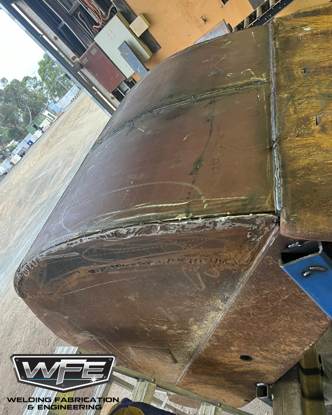 A large metal pipe with welds along its seams, placed on a worktable in a workshop, with electrical panels and wires in the background. The workshop has an open front, showing outdoor trees and vehicles.