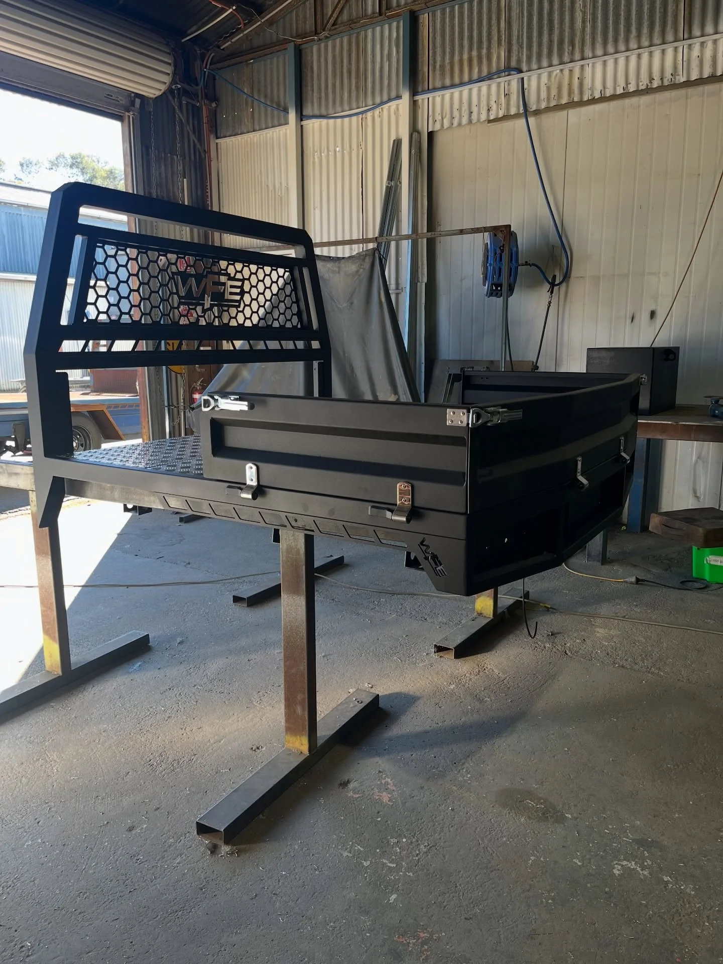 Inside a workshop, a black metal tool cart with a honeycomb pattern on its guard and a silver handle, placed on a black metal stand, with a workbench and some tools in the background.