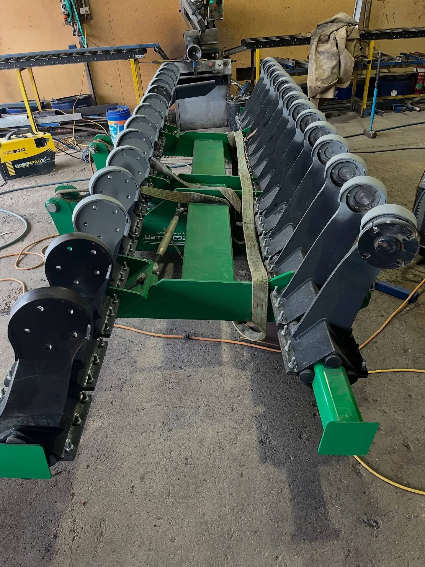 Green and black agricultural equipment with black wheels and straps inside a workshop with tools and equipment in background.