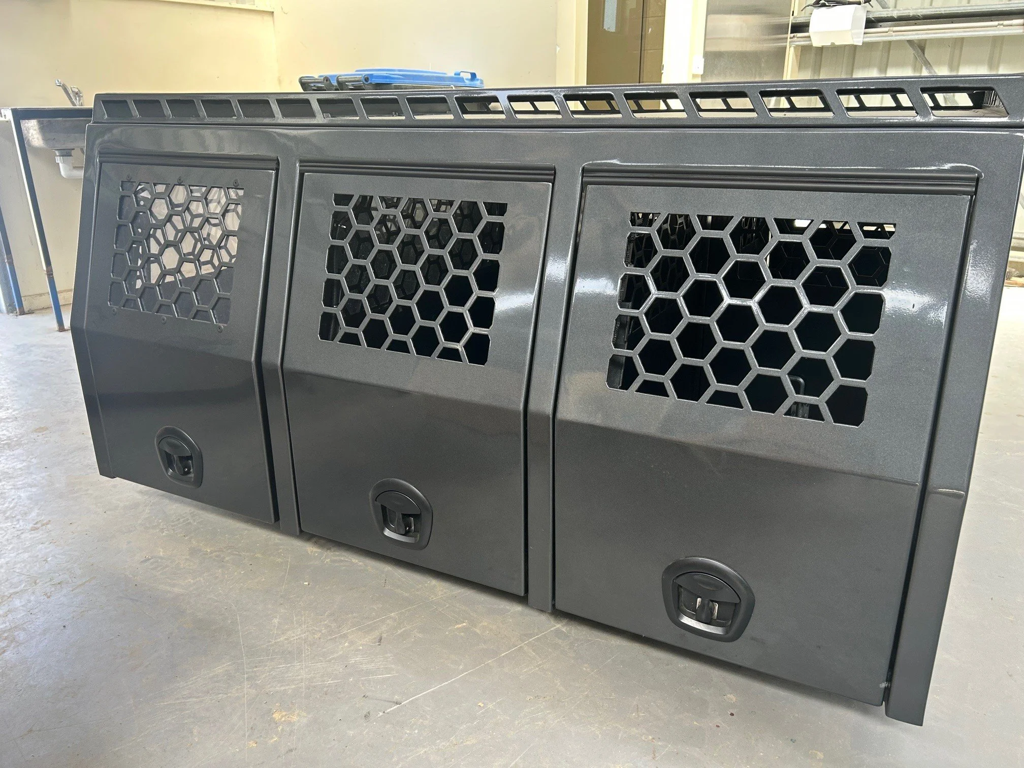 A black metal equipment or storage unit with honeycomb-patterned vents on its doors, situated in an industrial or workshop setting. There are blue-handled tools or objects on top of the unit, and a utility sink is visible in the background.