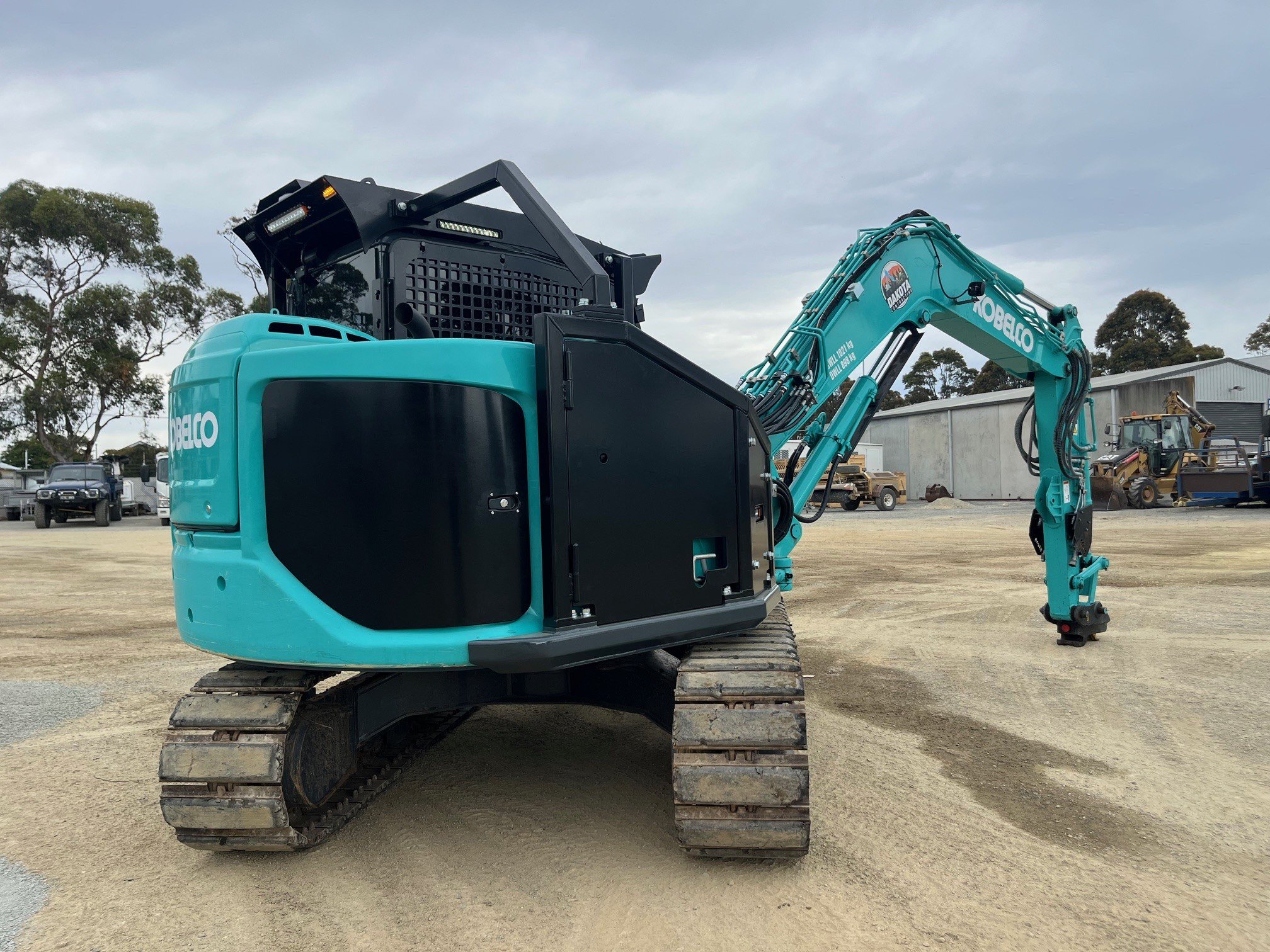 A teal-colored Kobelco compact excavator with black tracks, parked on a dirt lot with trees and industrial buildings in the background.