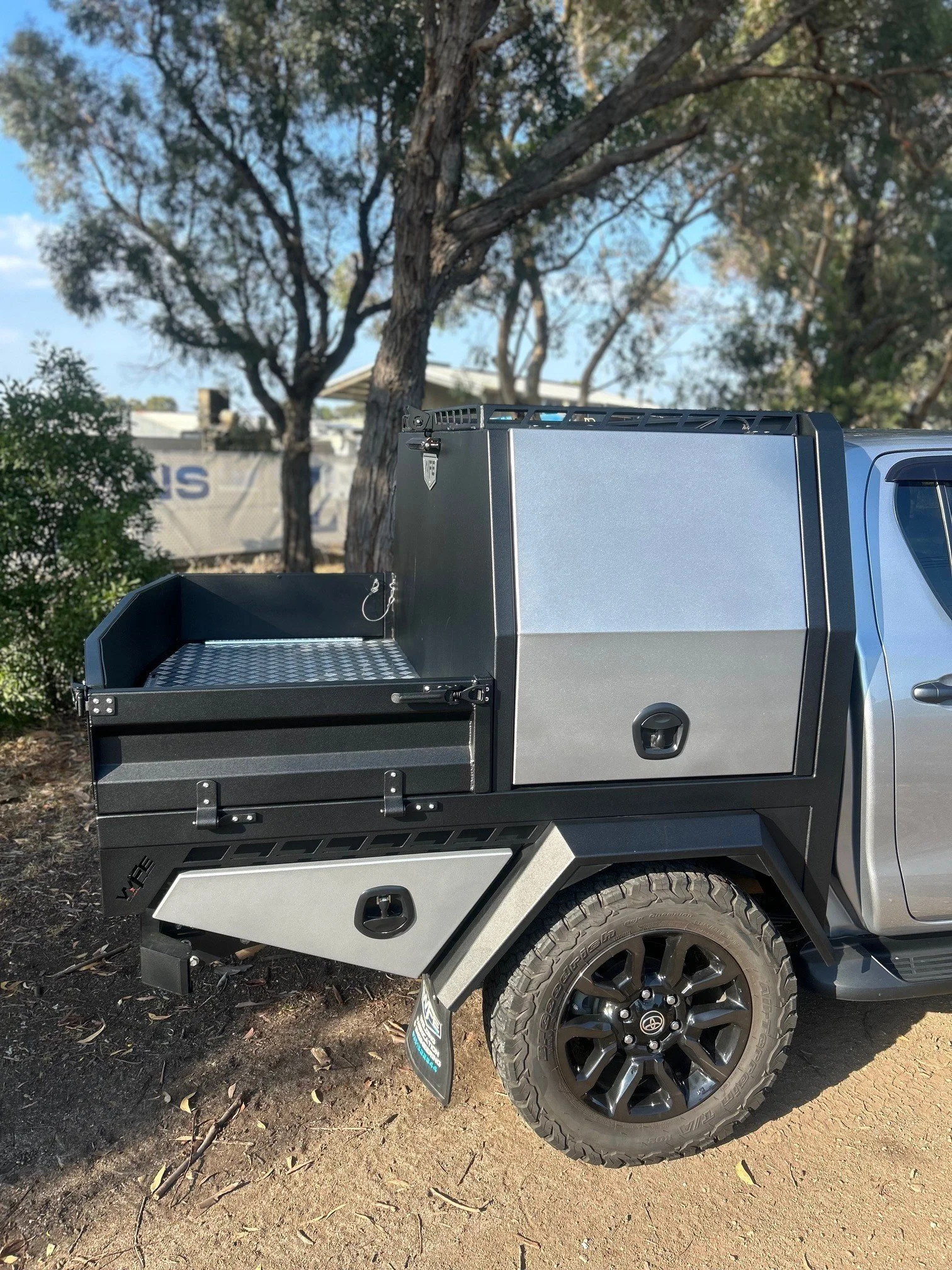 Close-up of a pickup truck's flatbed with storage compartments, black rims, and off-road tires, parked on dirt with trees and a fence in the background.