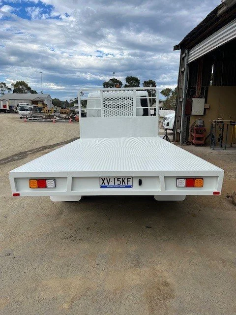 White flatbed utility truck with license plate XV 15KF parked on a dirt surface outside a workshop.