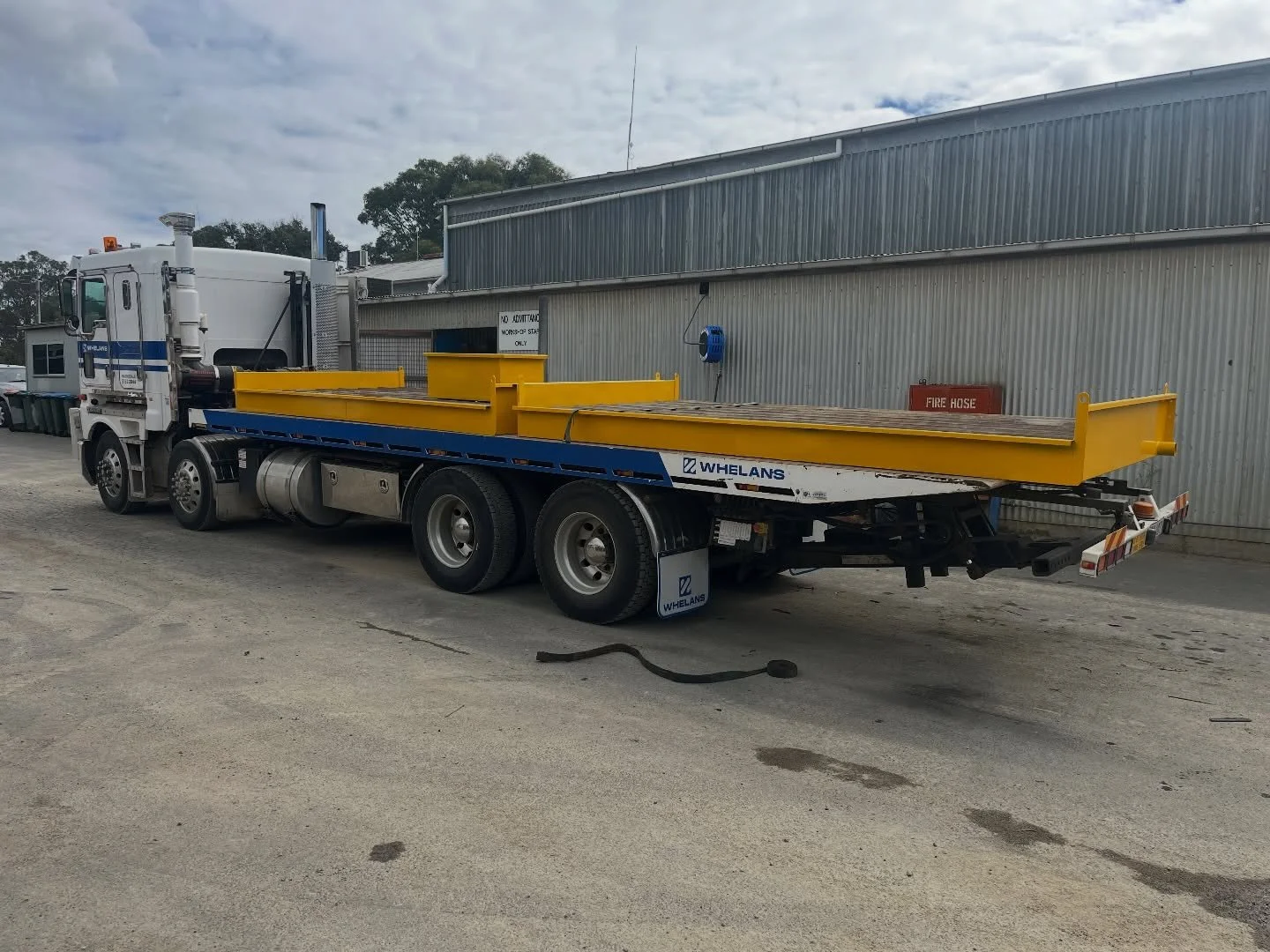 A flatbed tow truck with yellow rails and branding 'Whelans', parked on a concrete ground next to a metal building, under a cloudy sky.