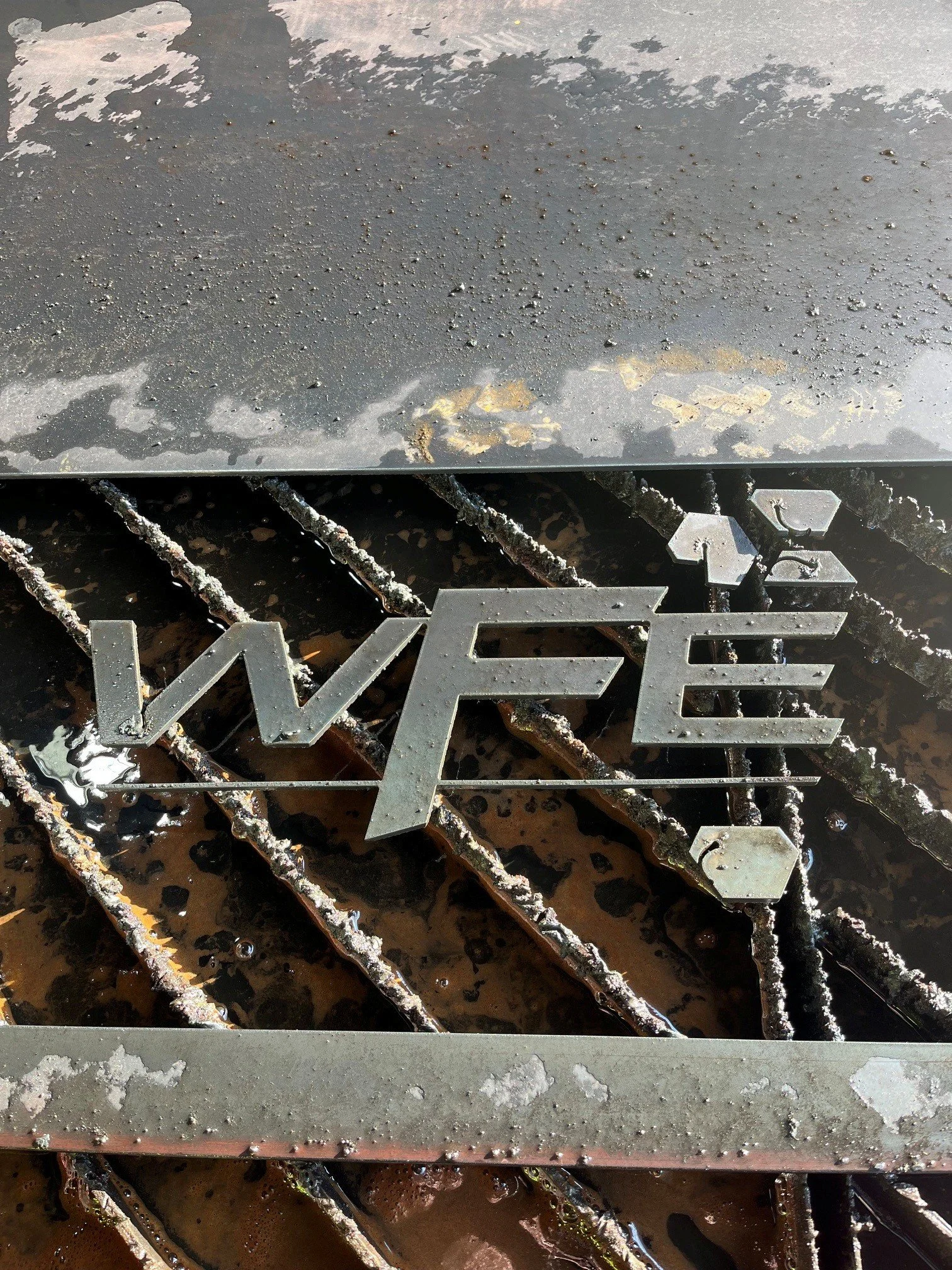Close-up of a black metal surface with some water droplets and dirt, part of a decorative metal structure with geometric shapes, and some rusted bar elements beneath.