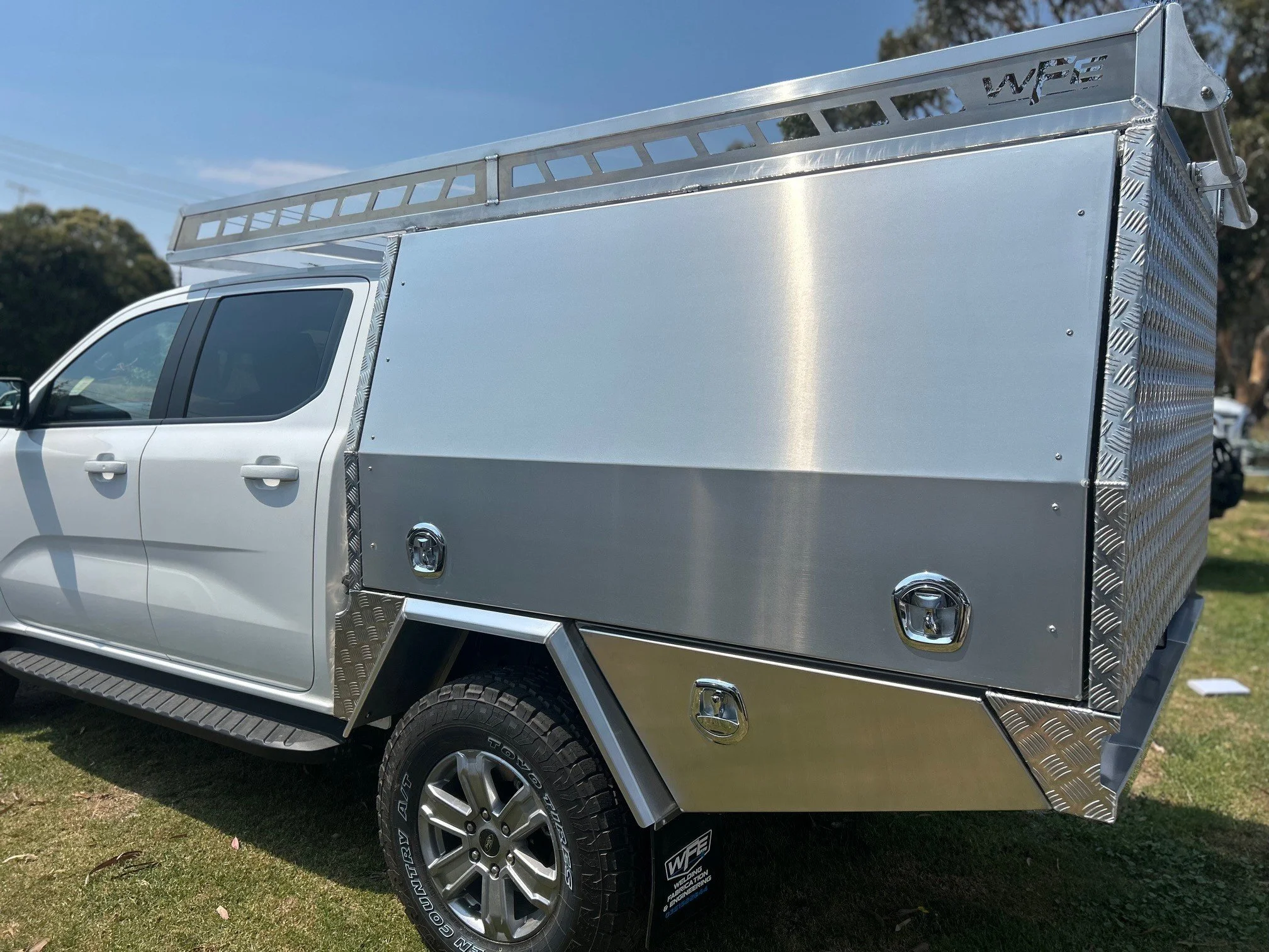 Silver pickup truck with a custom aluminum utility box on the bed, parked on grass with trees and a clear blue sky in the background.