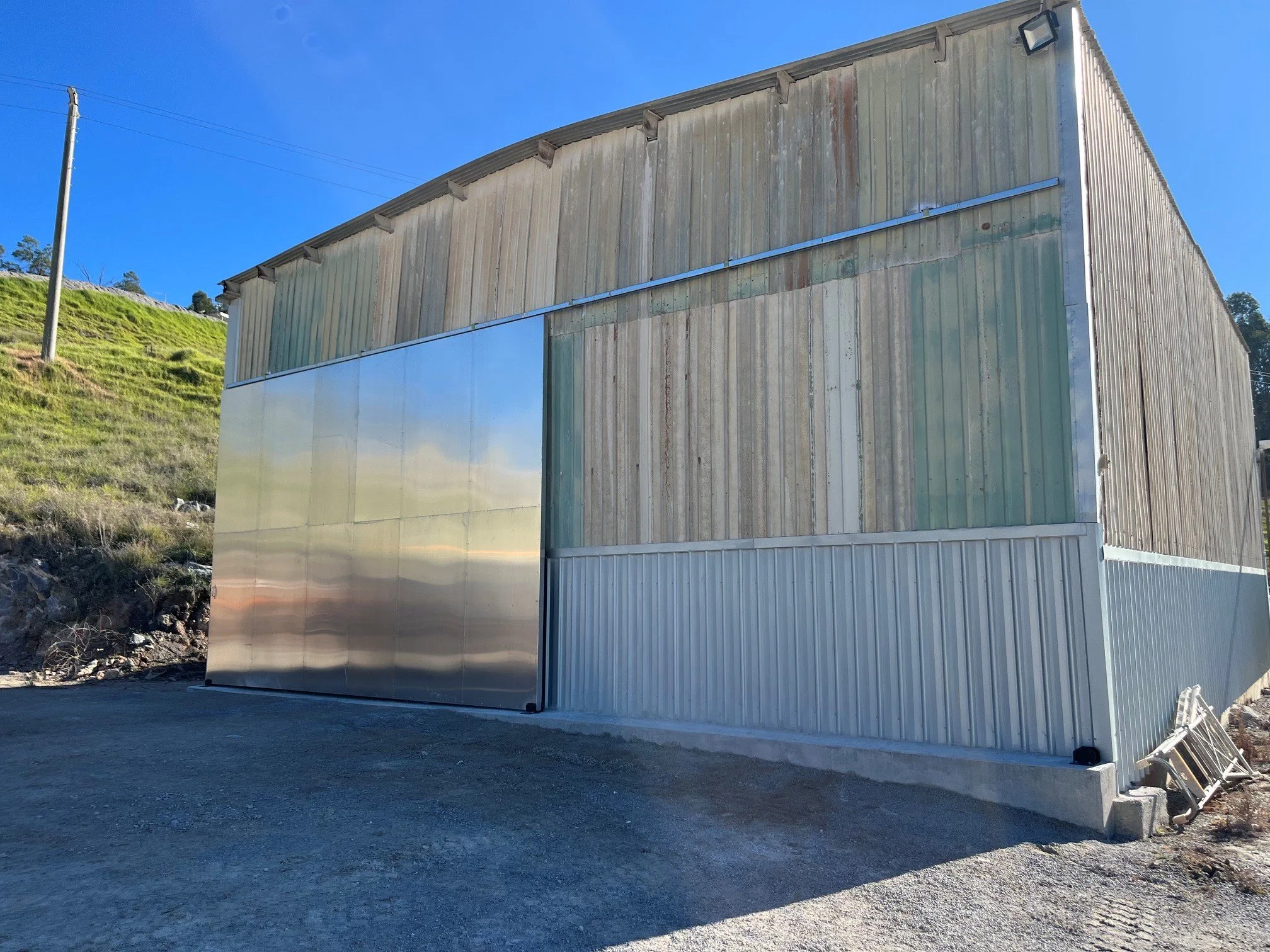 A modern building with metal siding, with the lower half made of reflective metal panels and the upper half of weathered vertical wooden planks, situated on a gravel lot with a hill and some trees in the background, under a clear blue sky.
