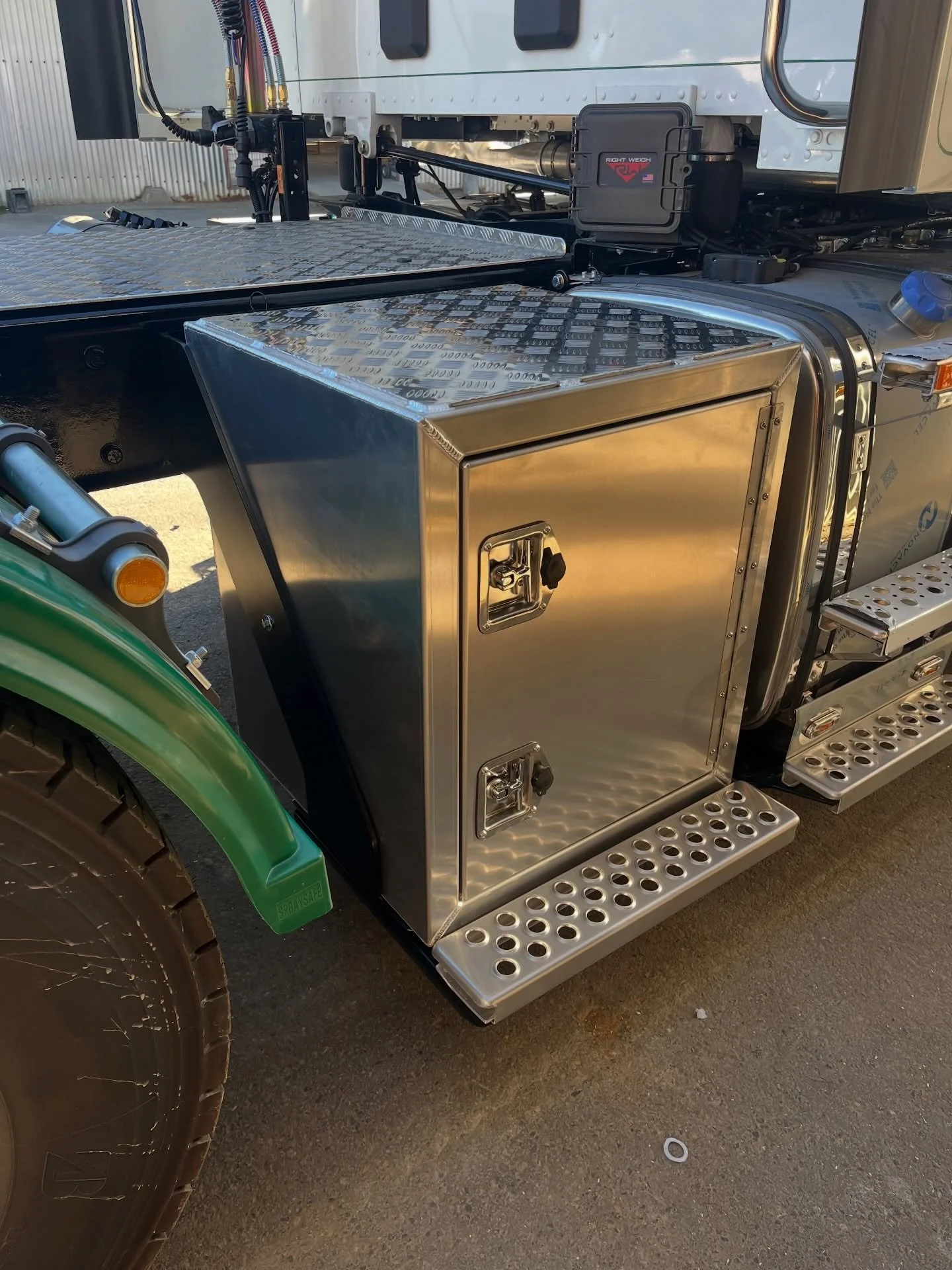 Close-up of a large truck's side, showing shiny metal storage compartments, a metal step, and a green wheel with a tire. Part of the truck's white and green exterior is visible.