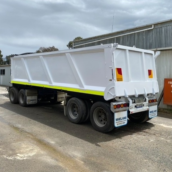 White dump truck with dual axles and large wheels parked on an unpaved surface outside a building with a cloudy sky overhead.