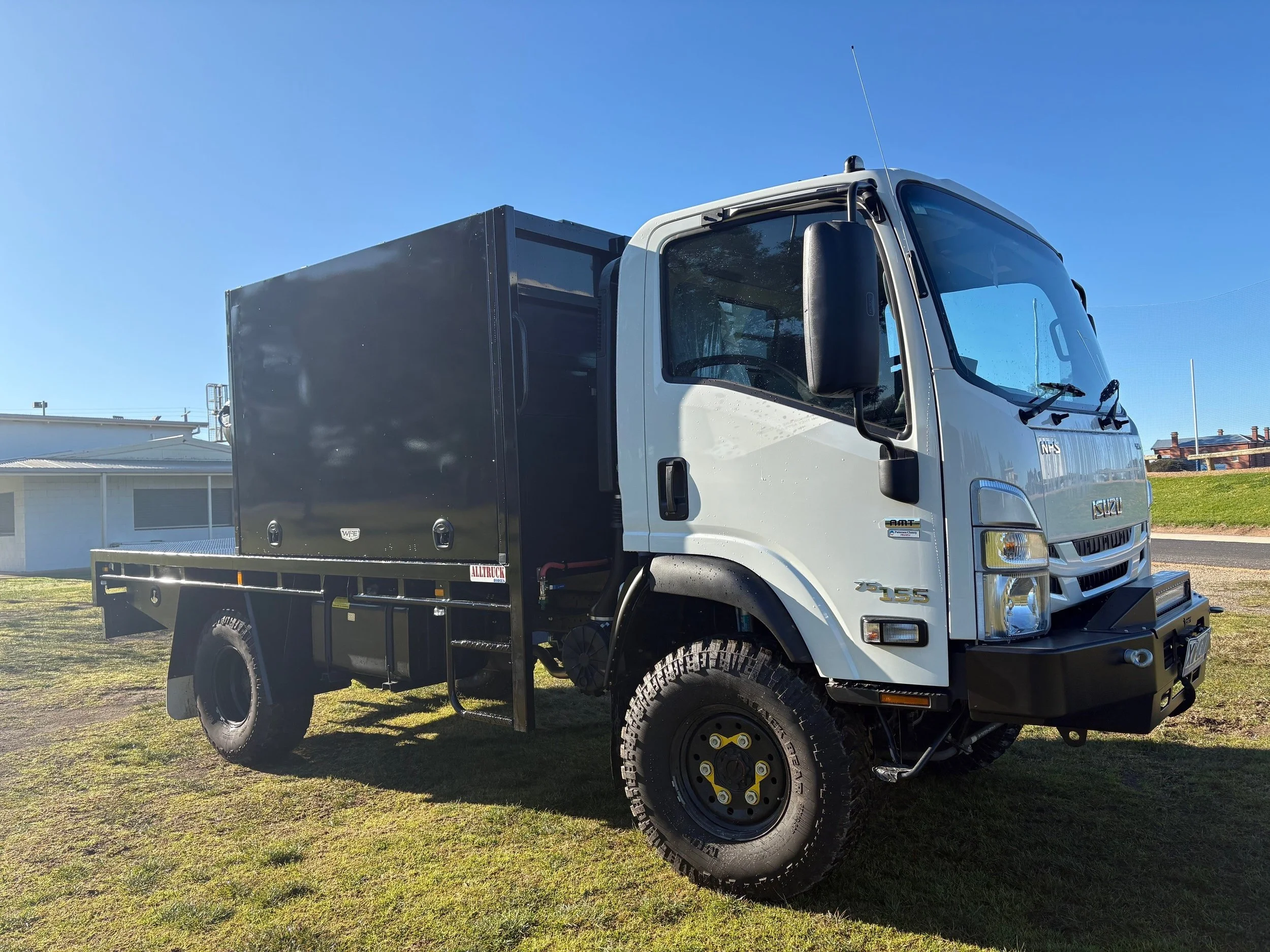 A white truck with a black cargo container parked on grass with clear blue sky and houses in the background.
