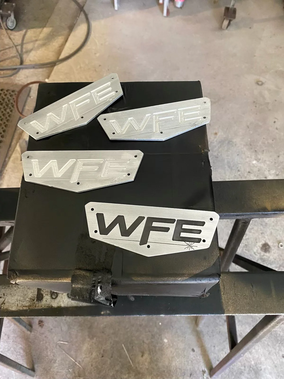 Metal signs with the text 'WFE' in bold letters, some with a stylized font, placed on a dark work surface in a workshop setting.