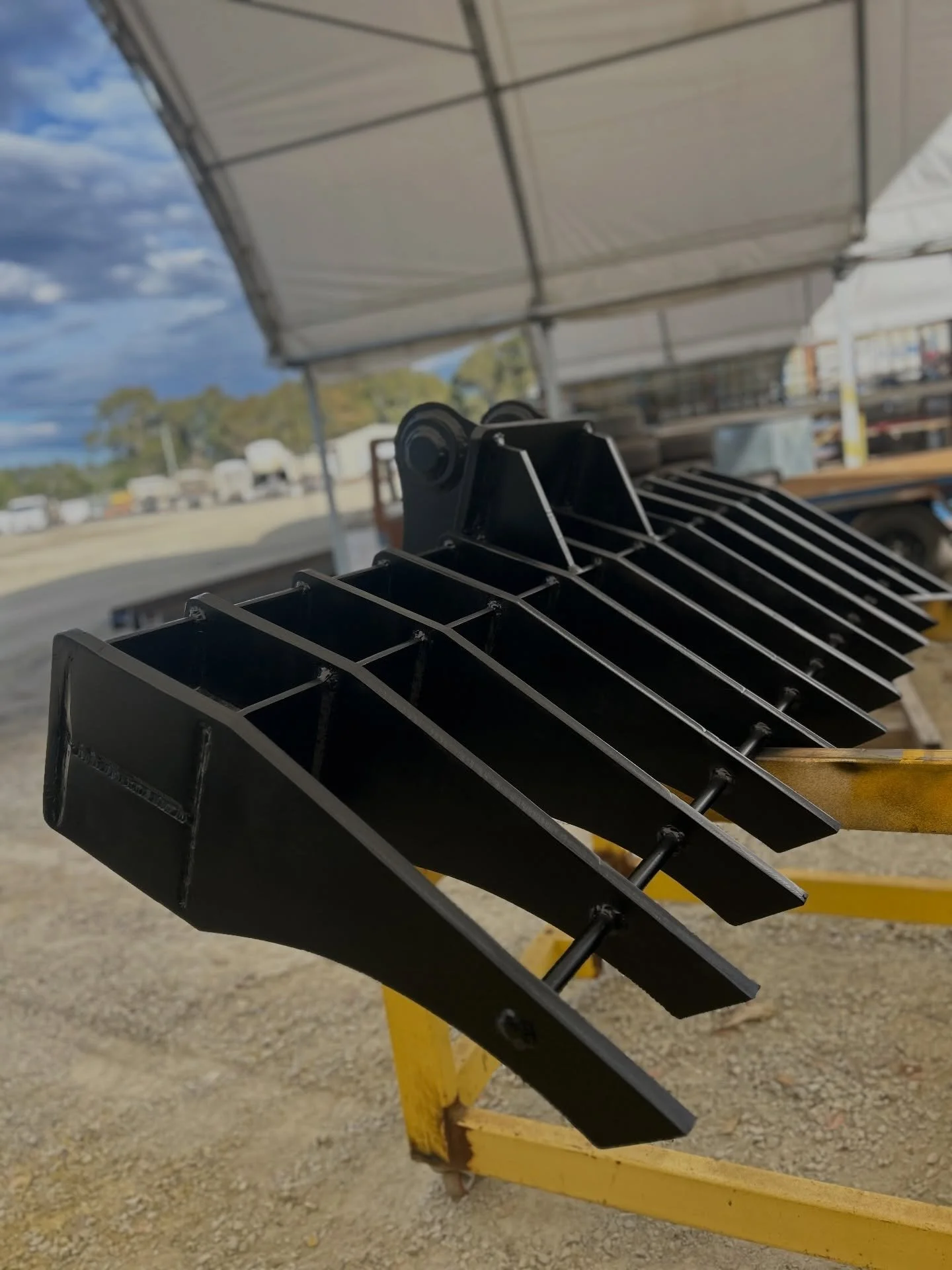 Black metal blades on a yellow metal stand outdoors under a white canopy with a partly cloudy sky in the background.