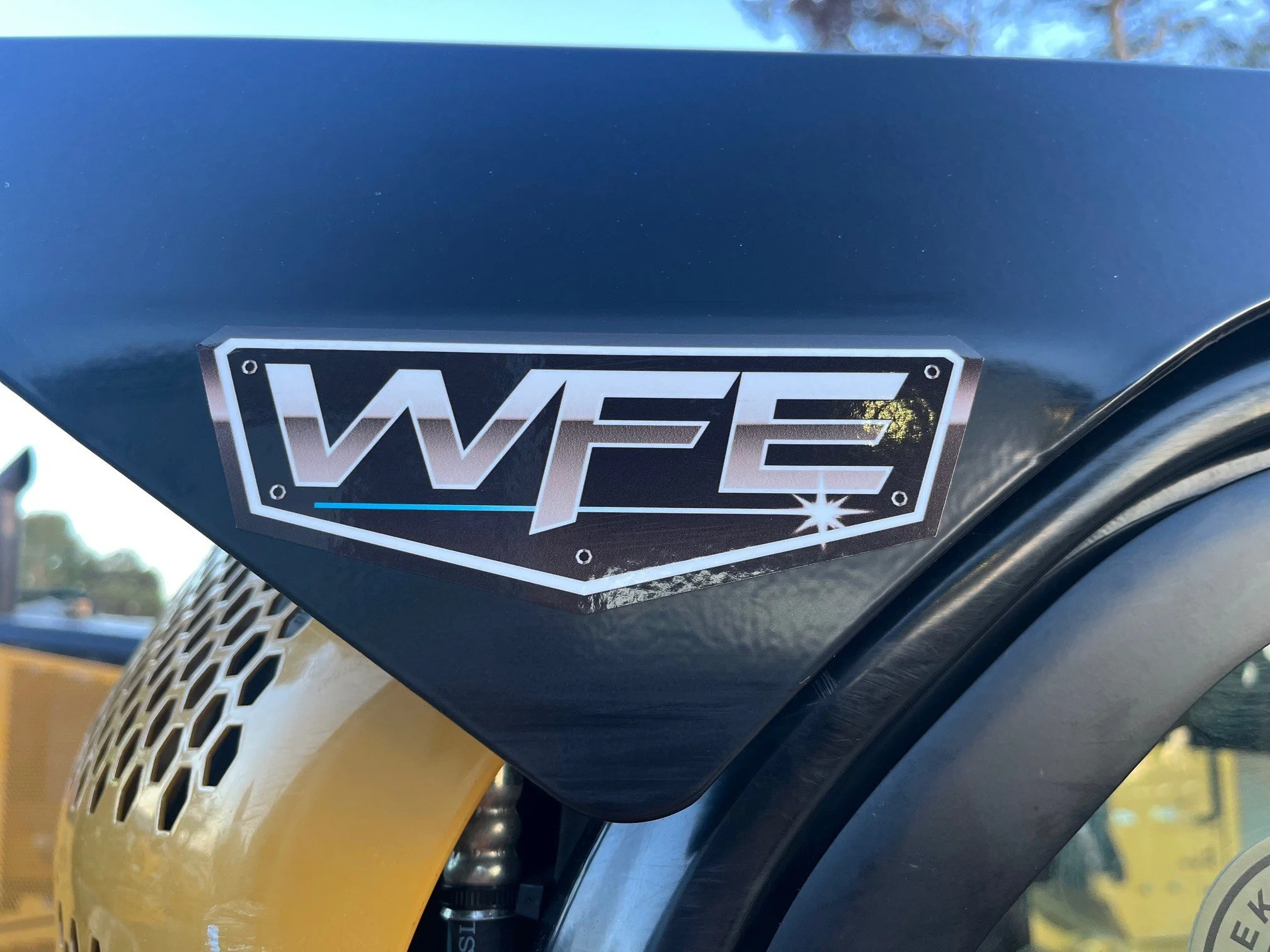 Close-up of motorcycle with a black and white sticker saying 'WEE' on the front. The motorcycle has a yellow front fender and tire with a black rim. In the background, there are trees and a blue sky.