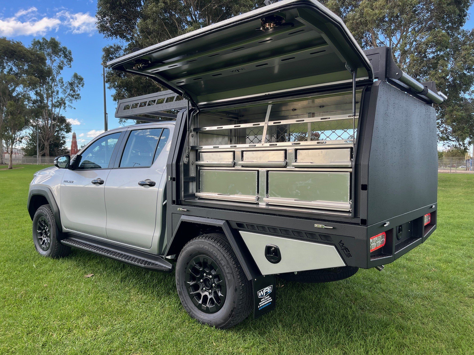 A white pickup truck with a black utility canopy, open back storage compartments, and an elevated roof rack, parked on a grassy field with trees and a partly cloudy sky in the background.
