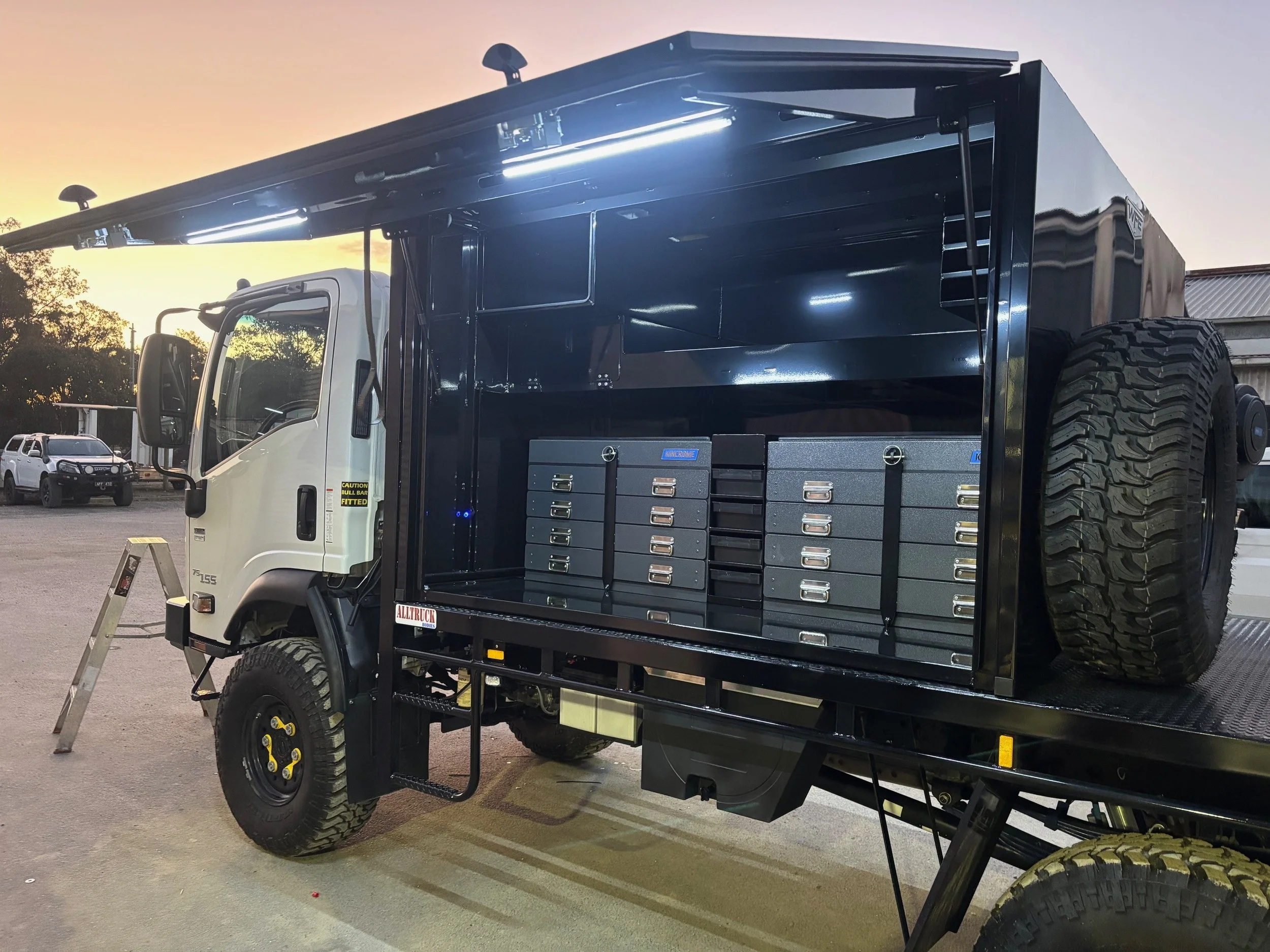 A black utility truck with built-in storage drawers and equipment compartments parked outdoors at sunset.