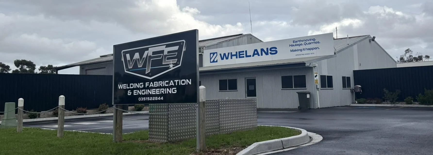 Exterior of a commercial building with signs for WFE Welding Fabrication & Engineering and Whelans, on a cloudy day. Parking lot in front with a small landscaped area.
