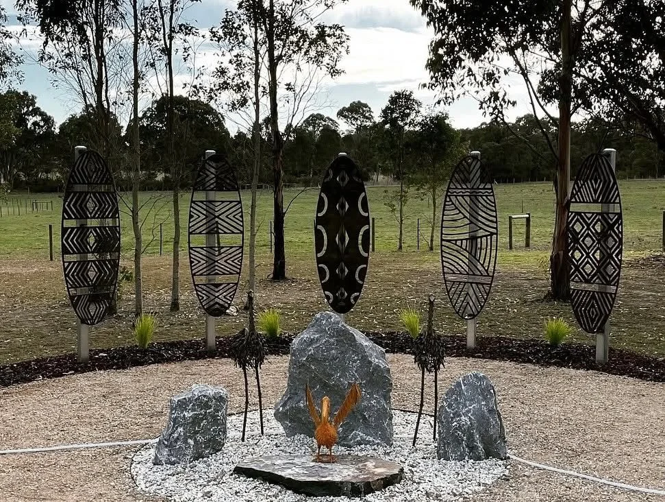 Outdoor sculpture with five tall, black tribal-style masks on poles, and a water feature with large rocks and a metal rooster in the center, surrounded by trees and grassy fields.