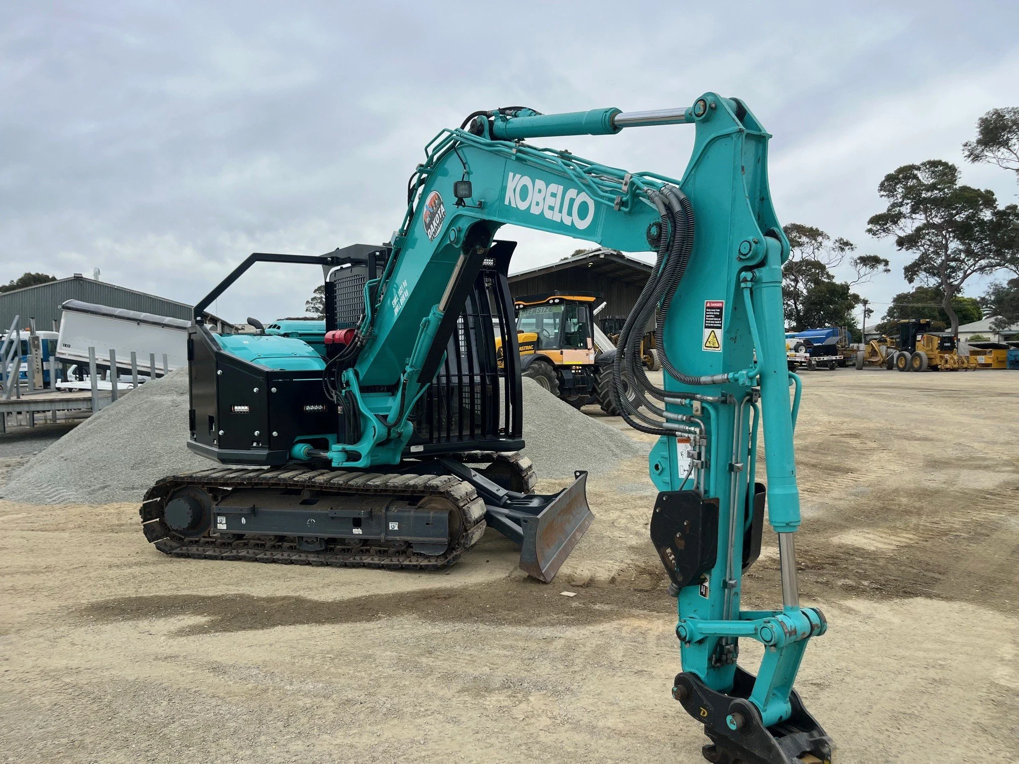 A Kobelco compact excavator with a teal arm and black tracks on a construction site, with various construction vehicles and equipment in the background under cloudy skies.