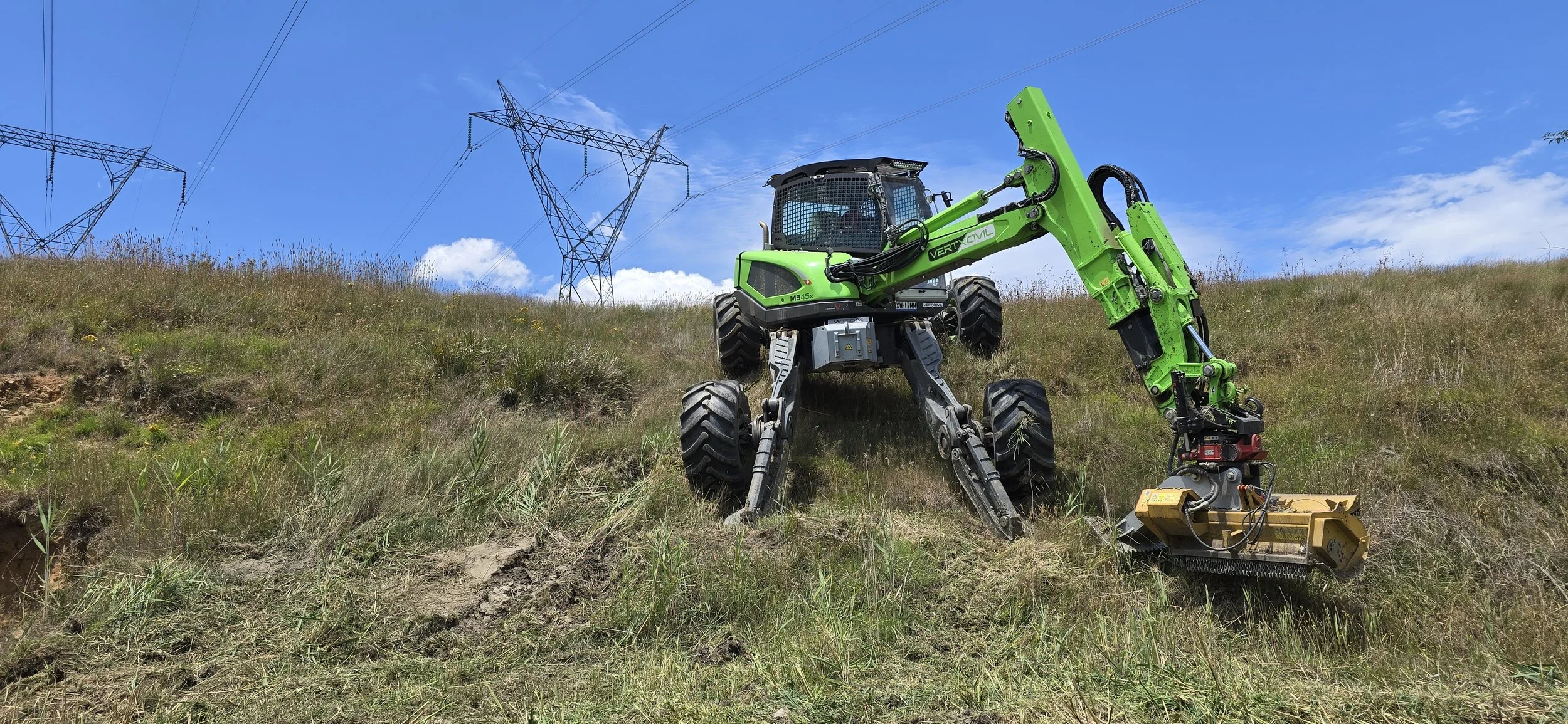 A green robotic piece of equipment working on grassland with power line towers in the background under a blue sky.