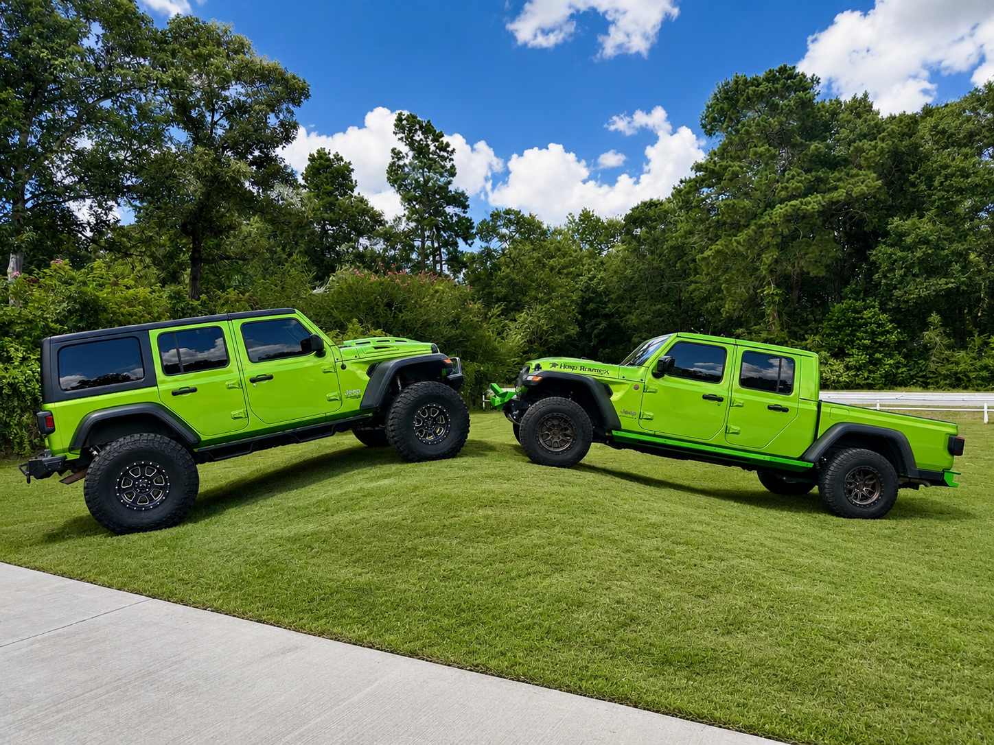 Two bright green Jeep vehicles with large tires parked on a grassy area, facing each other, under a blue sky with clouds, surrounded by trees.