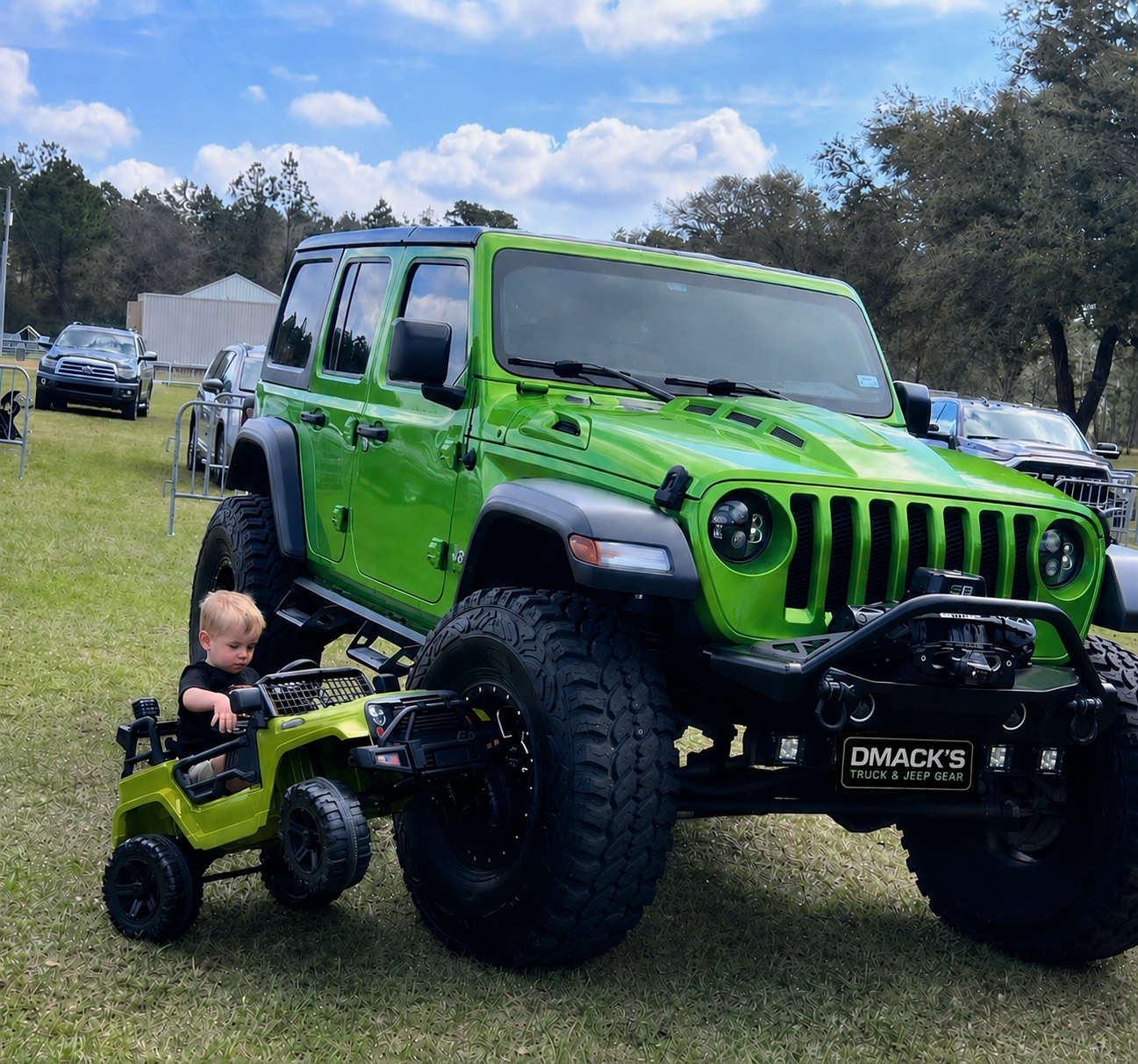A young boy sitting in a small green toy off-road vehicle next to a large green Jeep Wrangler on a grassy field with other vehicles and trees in the background.