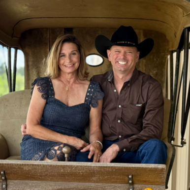 A woman and a man sitting together inside a vintage vehicle, both smiling. The woman has shoulder-length light brown hair and is wearing a dark blue dress with lace details. The man is wearing a black cowboy hat and a brown shirt.