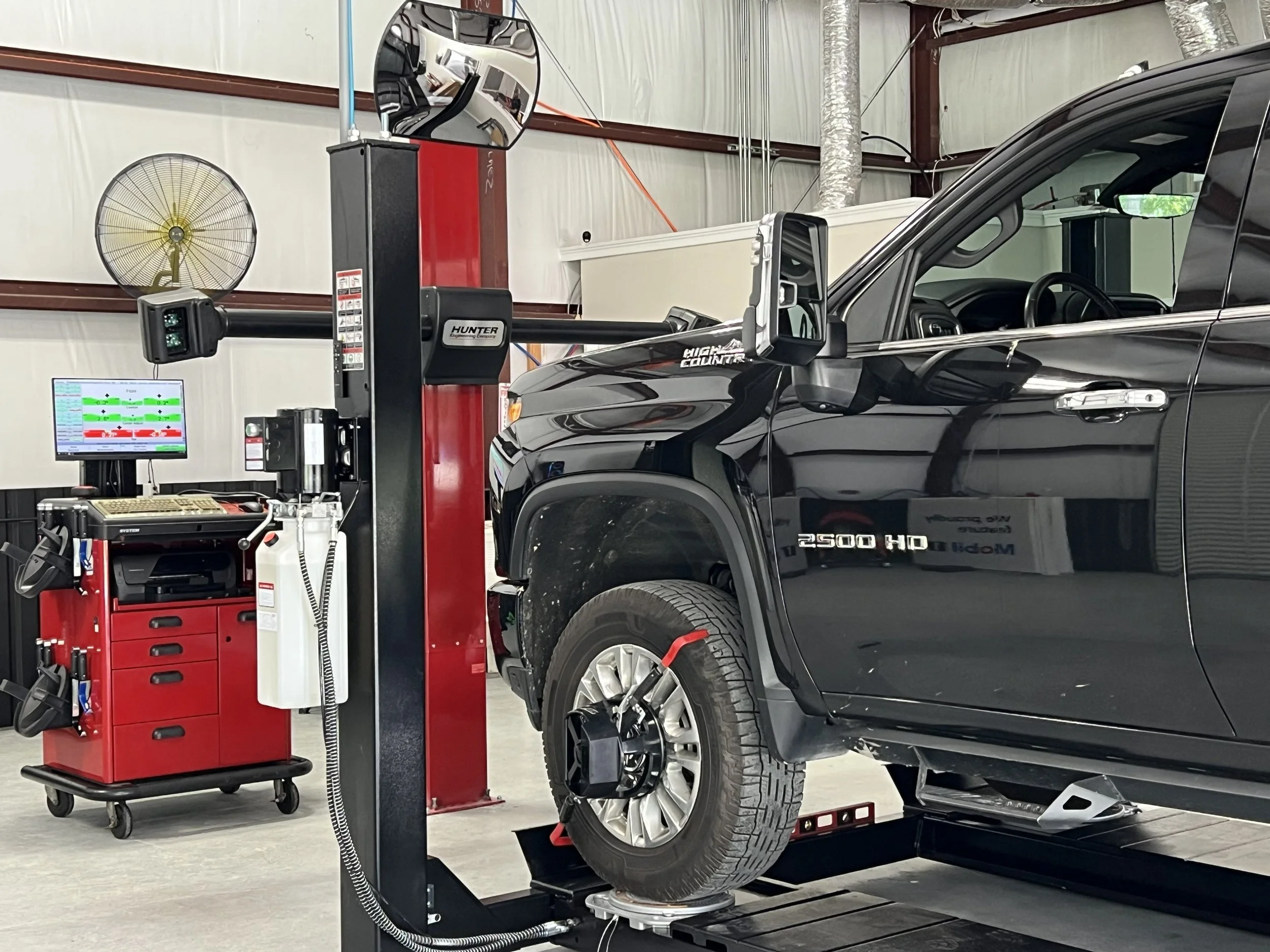 A black truck in an automotive shop, getting an alignment, with a tool cart nearby, a fan, a computer monitor, and various equipment in the background.