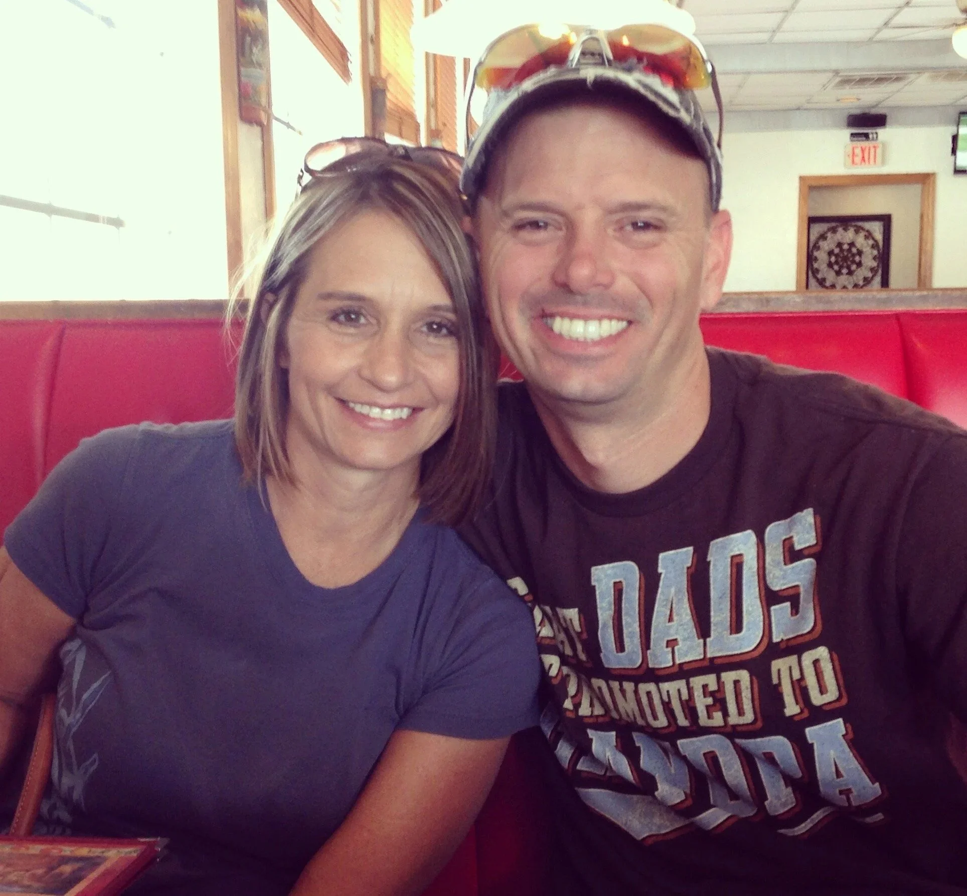A smiling woman with light brown hair and a man with a cap and sunglasses on his head sitting closely together in a restaurant booth.