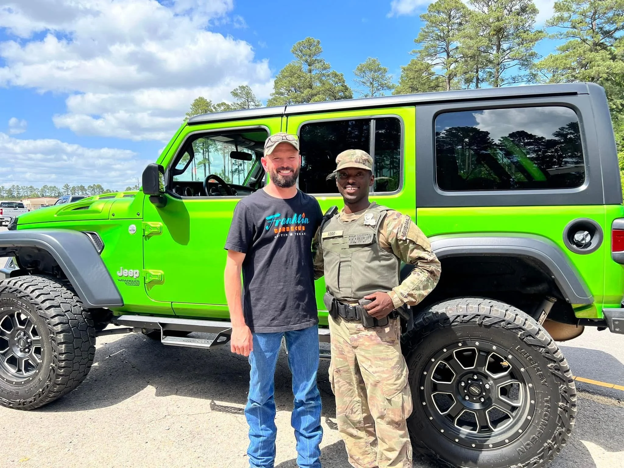 Two men standing in front of a bright green Jeep Wrangler Unlimited, smiling outdoors on a sunny day with a backdrop of trees and a blue sky with clouds.