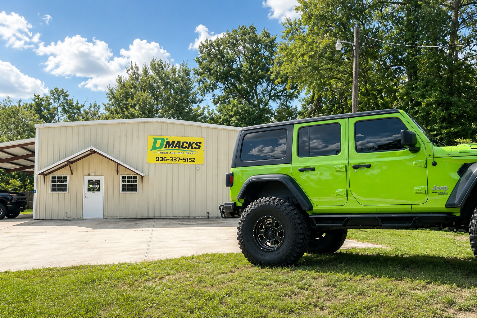 Green Jeep SUV parked in front of a beige building with a yellow sign that reads "D'Macks Truck and Jeep Gear." The building has a white door and two windows, with a small roof over the entrance. The scene is outdoors on a sunny day with blue sky and