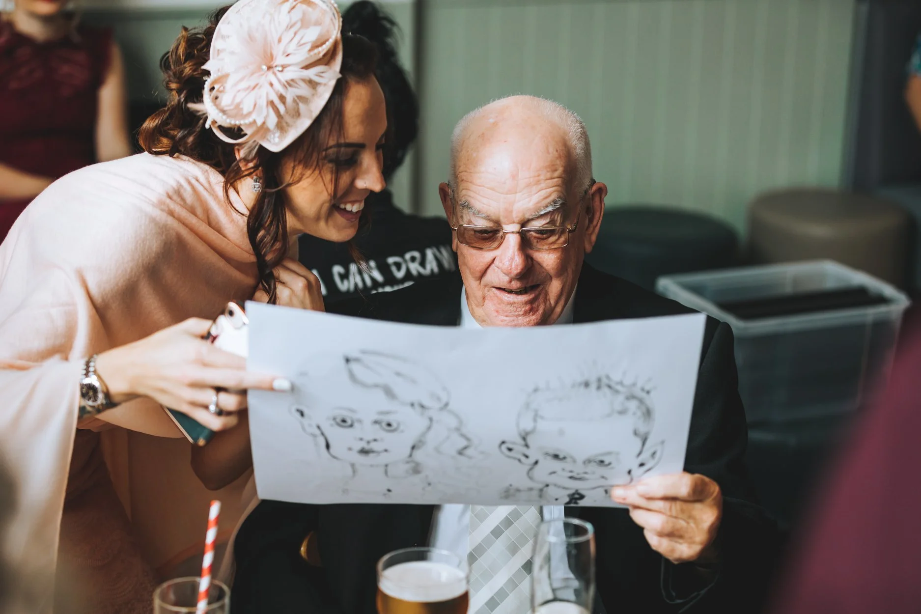 A woman with a large floral headpiece and a man with glasses looking at a drawing of faces, with drinks on the table.