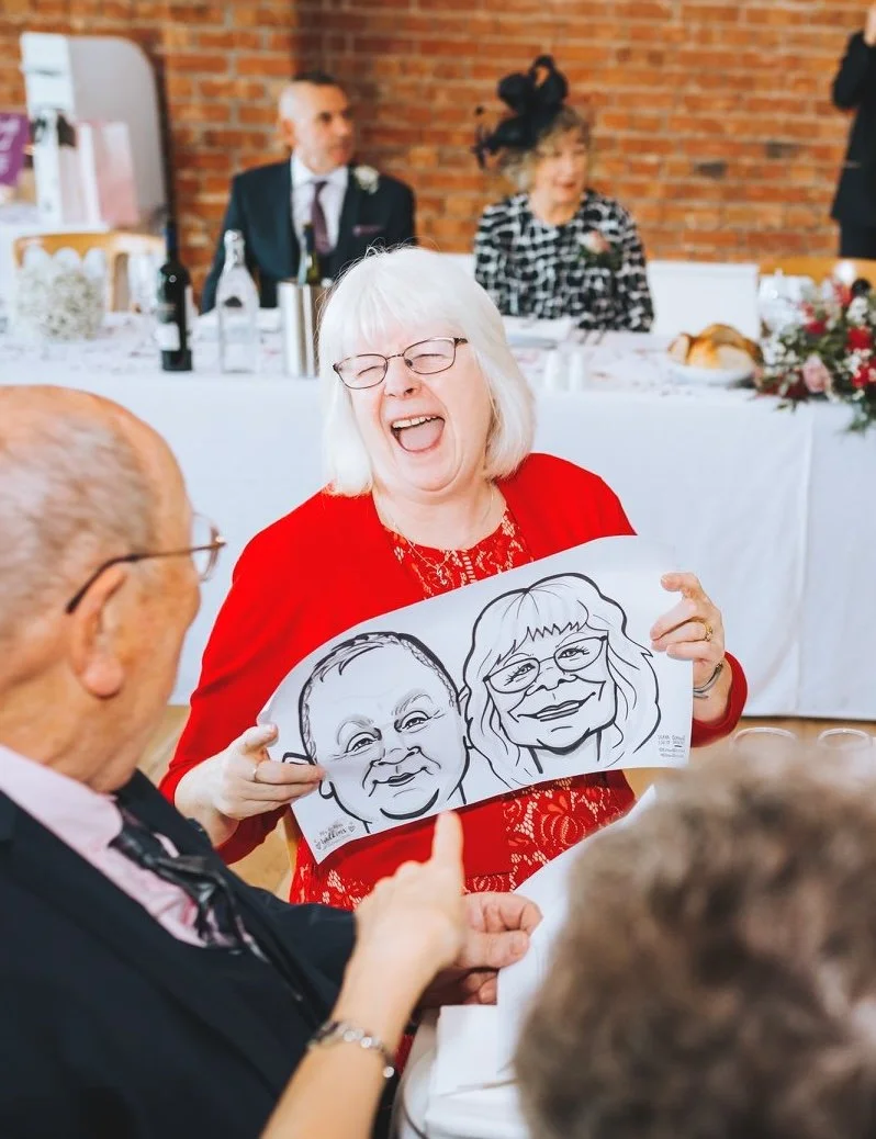 Woman holding a caricature of a man and woman, laughing joyfully at a wedding, with others seated at a decorated table in the background.
