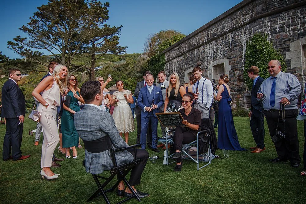 A group of people dressed in formal attire gathered outdoors around a couple seated in chairs, with some holding drinks and smiling. The setting appears to be a wedding or celebration on a sunny day, with a stone wall, trees, and grass in the backgro