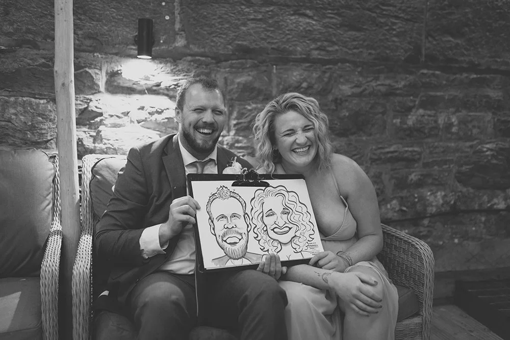 A man and woman sitting on a couch, holding a caricature drawing of themselves, both smiling and laughing in a cozy room with stone walls.