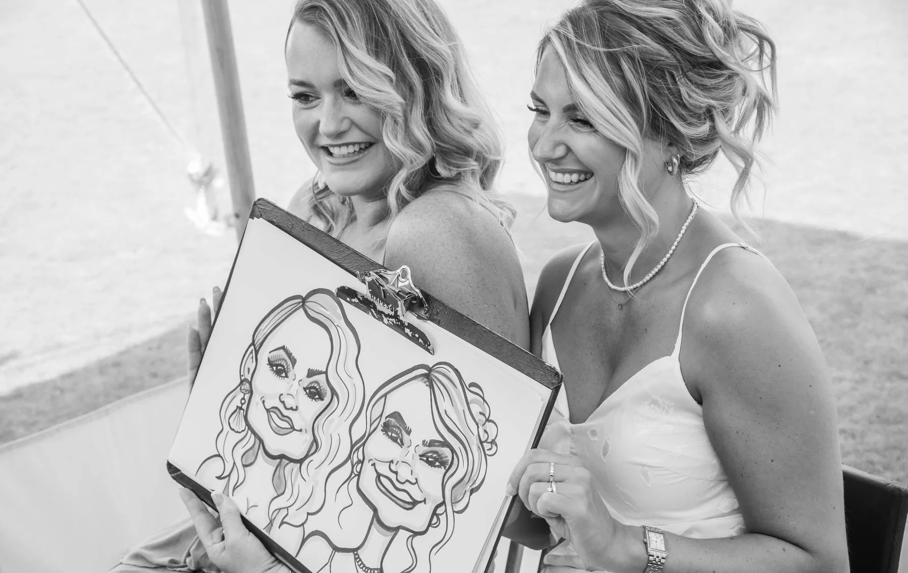 Two smiling women, dressed in white, are looking at caricature drawings of themselves on a clipboard, sitting in a marquee at a wedding.